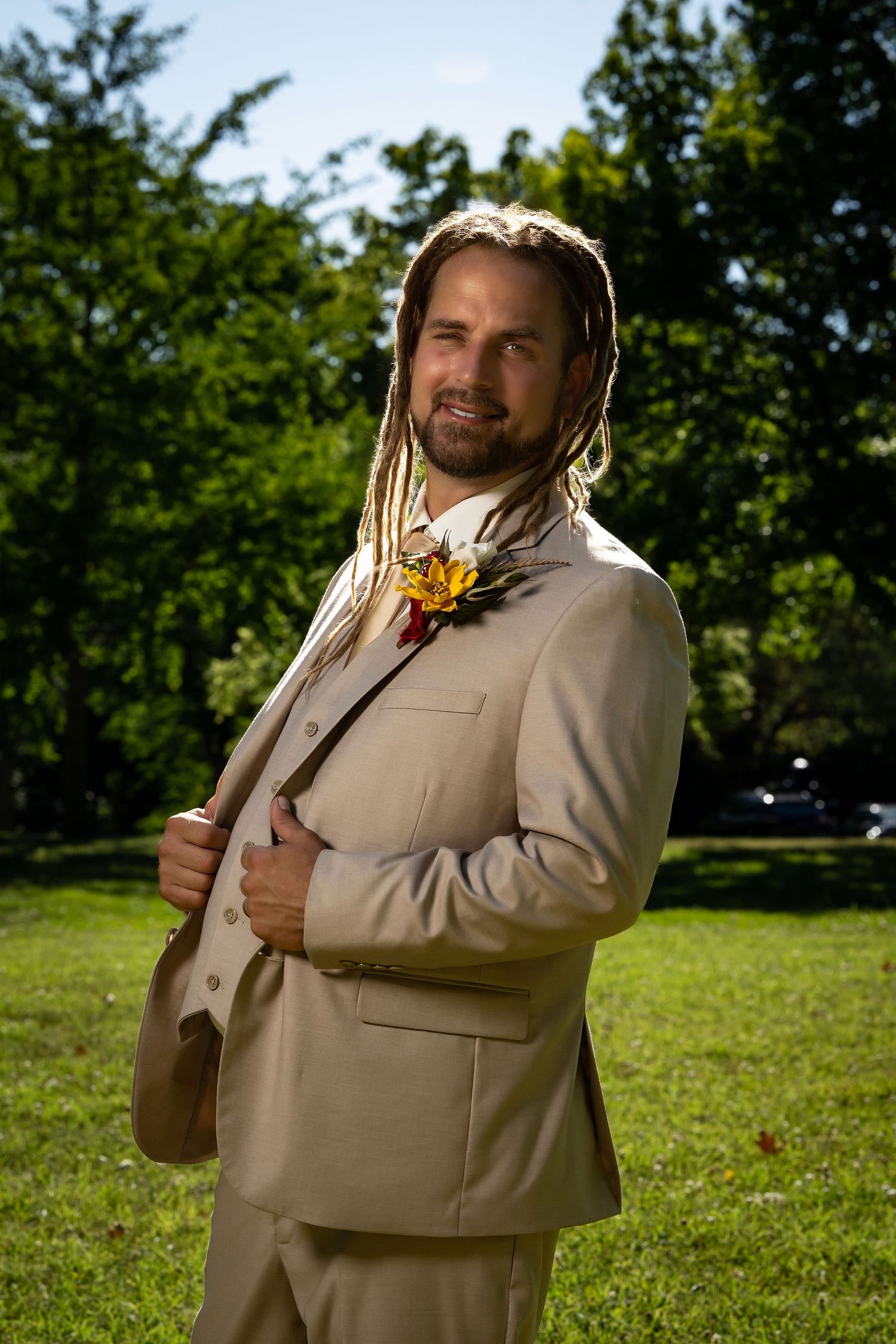 Groom in beige suit poses outdoors with floral boutonniere against green landscape.