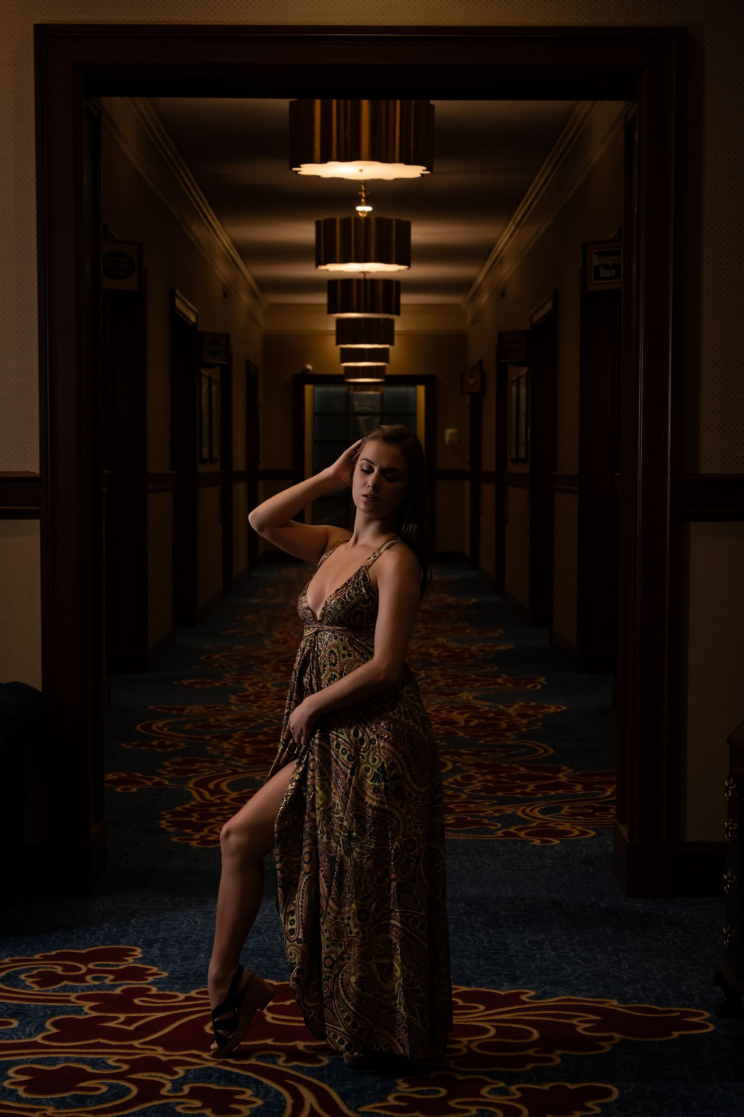 Glamorous portrait of model in gold dress posing in dimly lit hotel hallway.