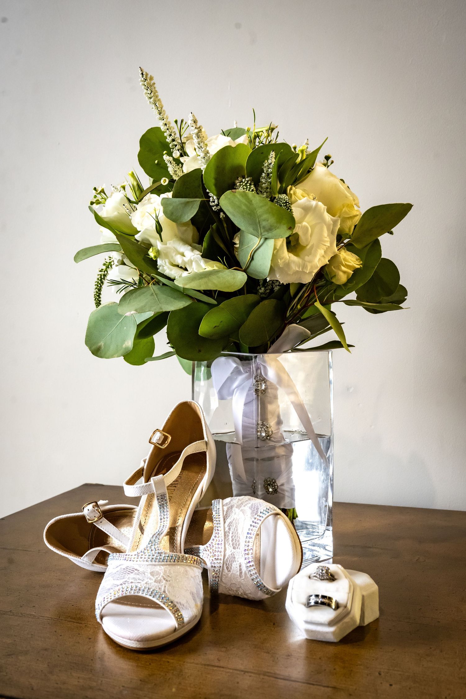 White bridal shoes displayed next to floral arrangements in silver vases on wooden table.