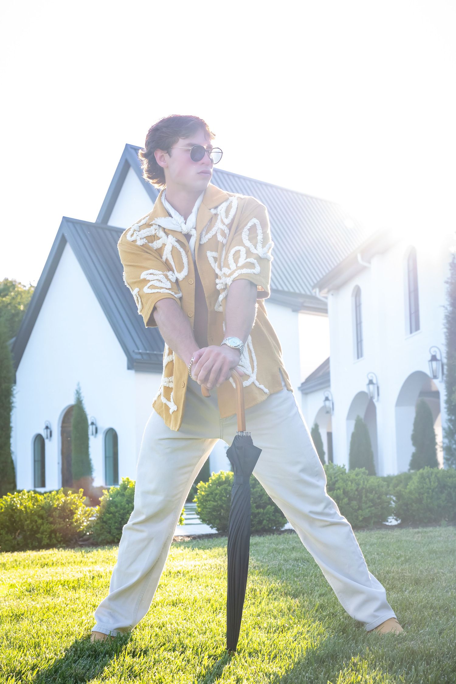 Person in yellow patterned shirt and white pants poses near white houses on sunny lawn.