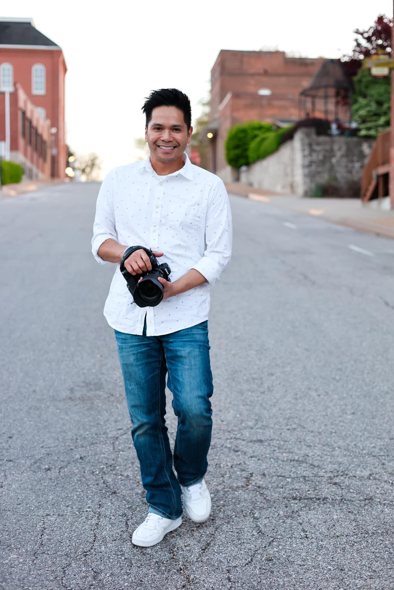 A smiling man in a white shirt and jeans holds a DSLR camera while standing on a street in an urban setting.