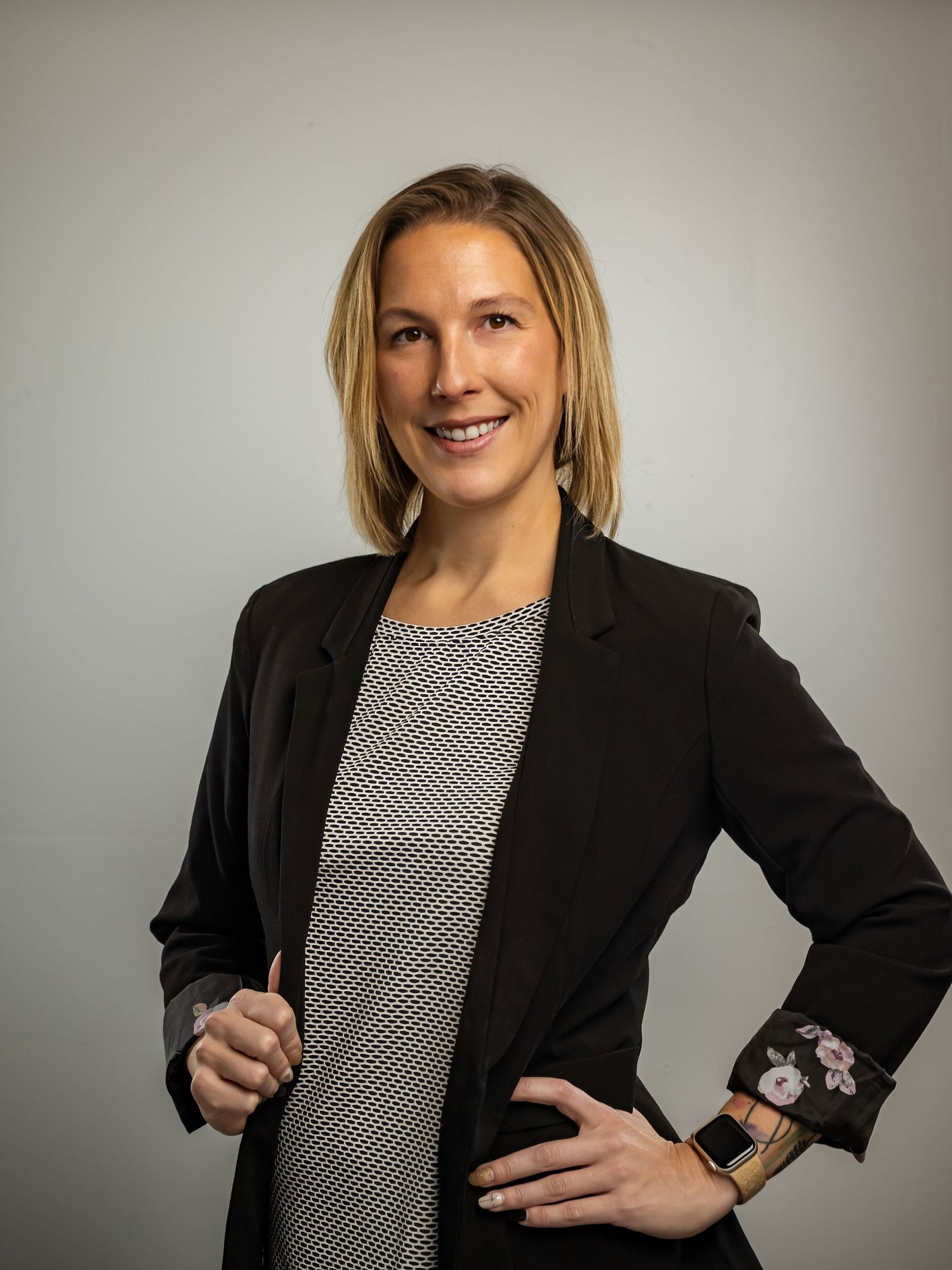 Professional woman in black blazer smiling confidently with hand on hip against neutral background.