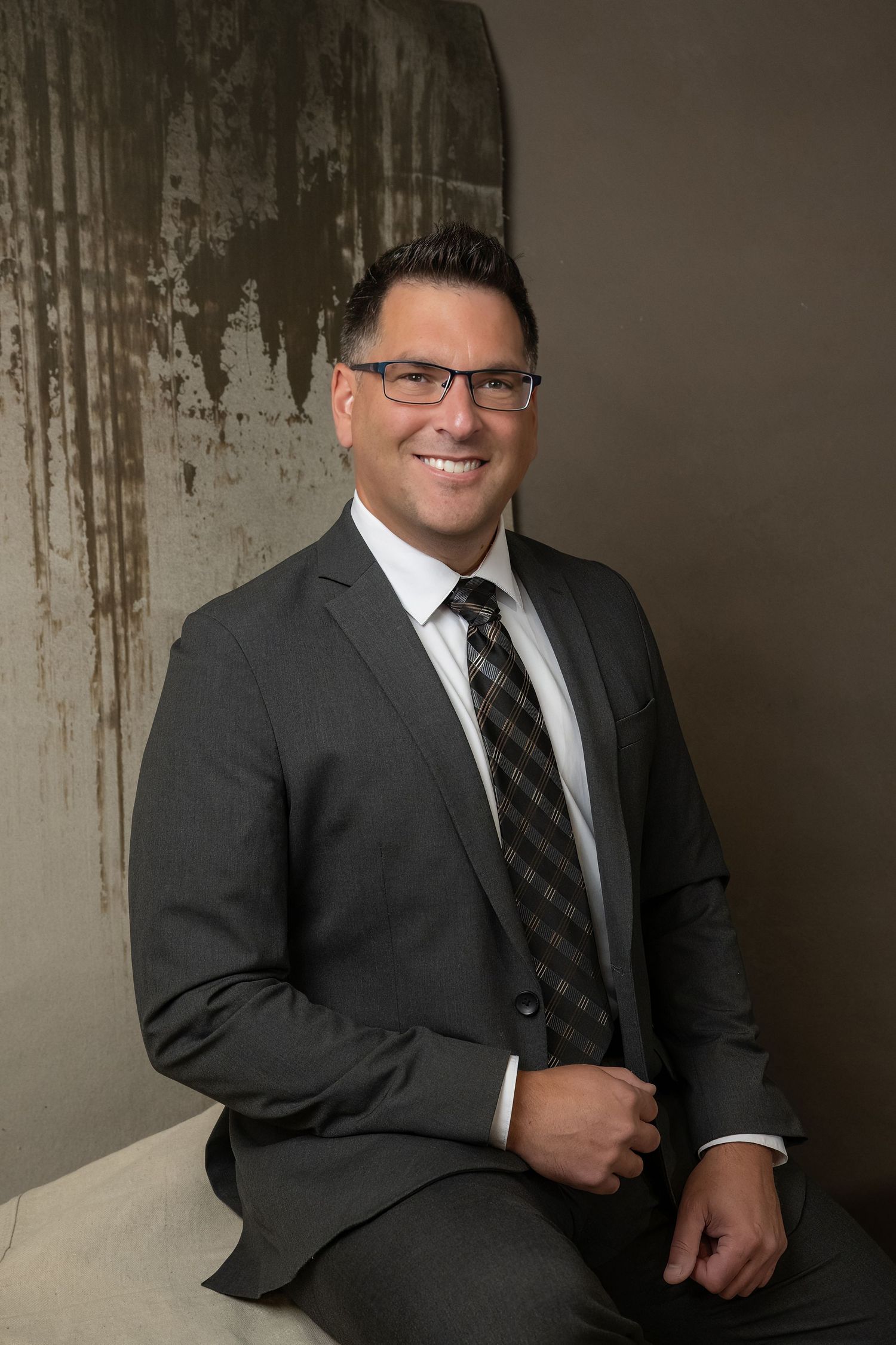 A smiling professional man in a gray suit and plaid tie, wearing glasses, poses confidently for a business portrait.