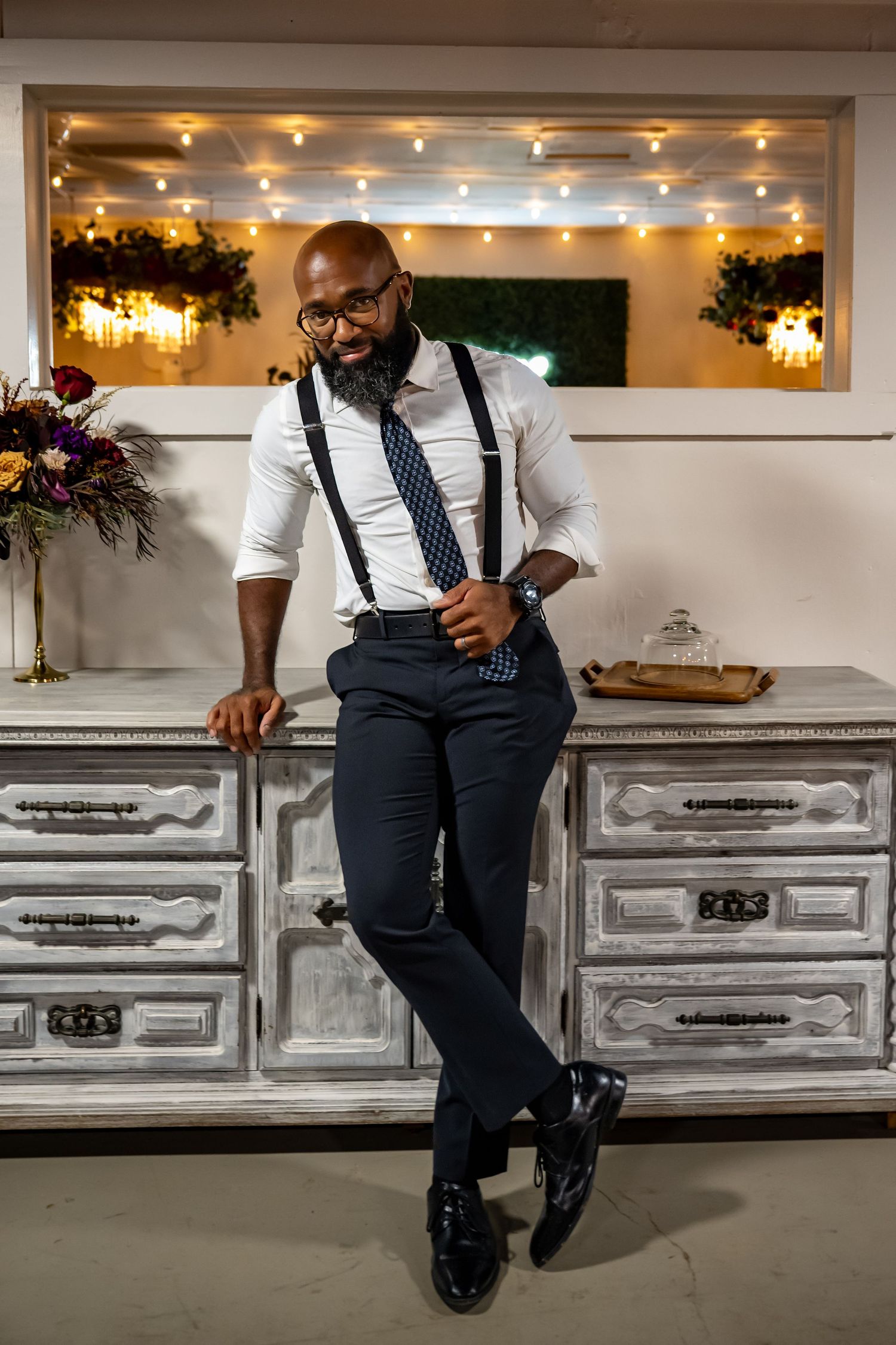 Stylish bald man with beard wearing suspenders, tie, and dress pants leaning against vintage dresser with string lights.
