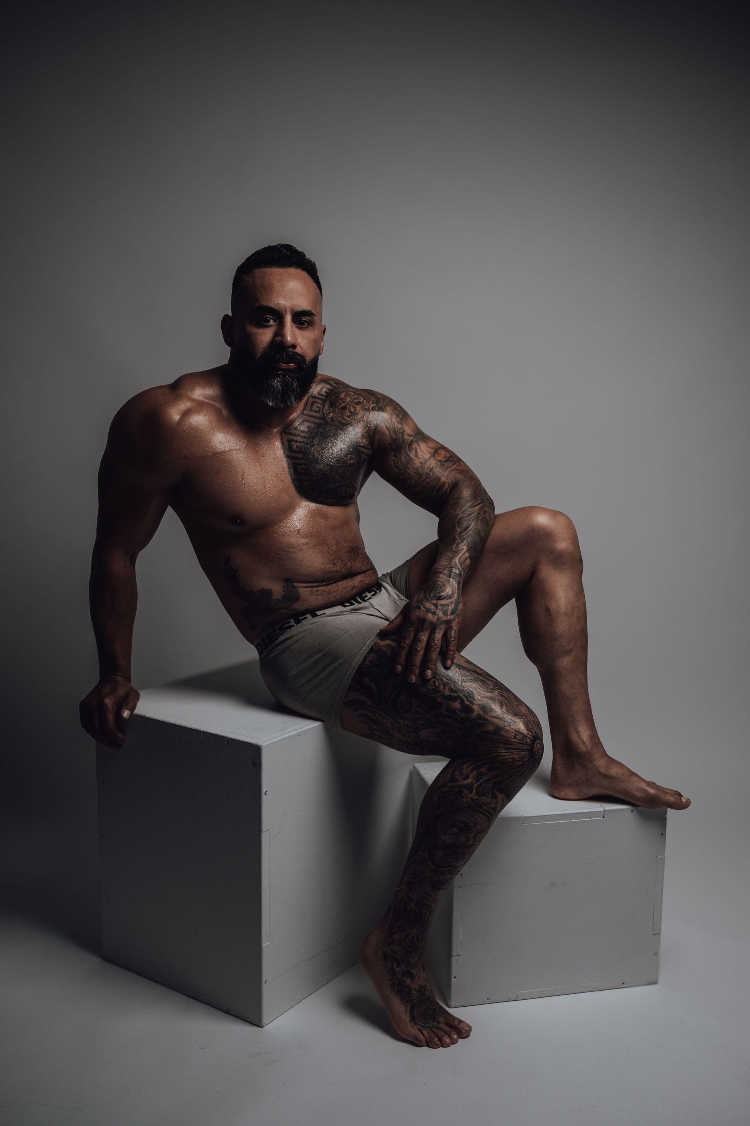 Muscular tattooed man with beard sitting shirtless on white cube in studio, wearing gray shorts.