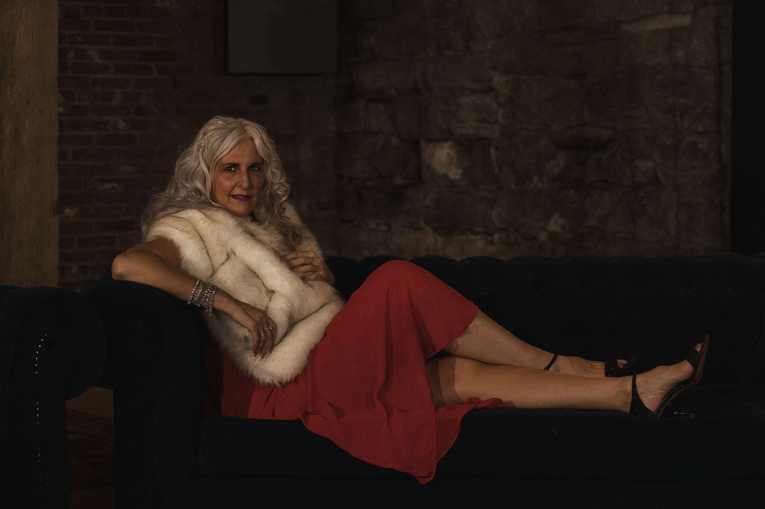 Elegant woman with silver hair reclining on dark sofa wearing red skirt and white fur stole in moody setting.