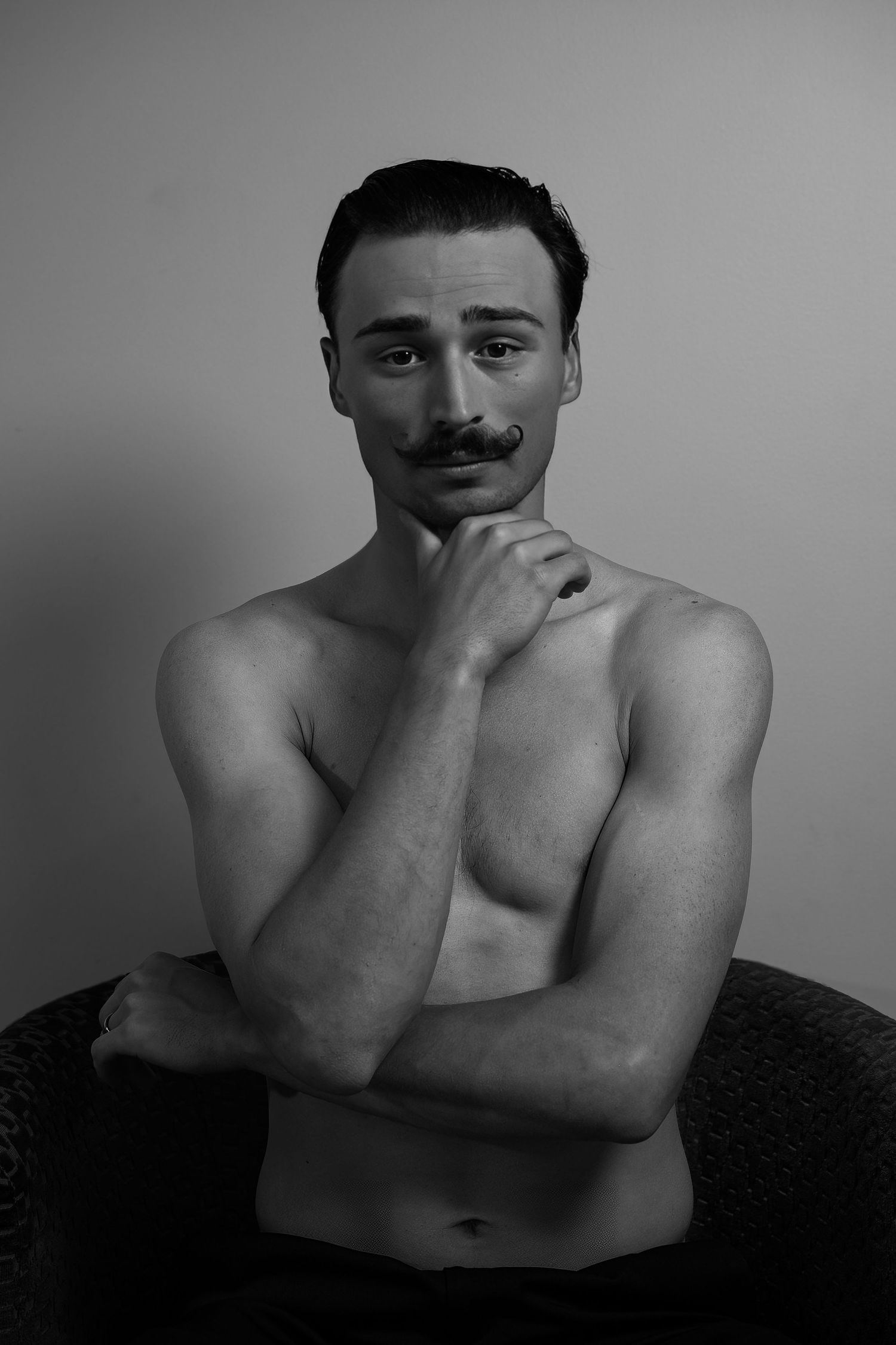 Black and white portrait of a shirtless man with a mustache posing thoughtfully with hand under chin.