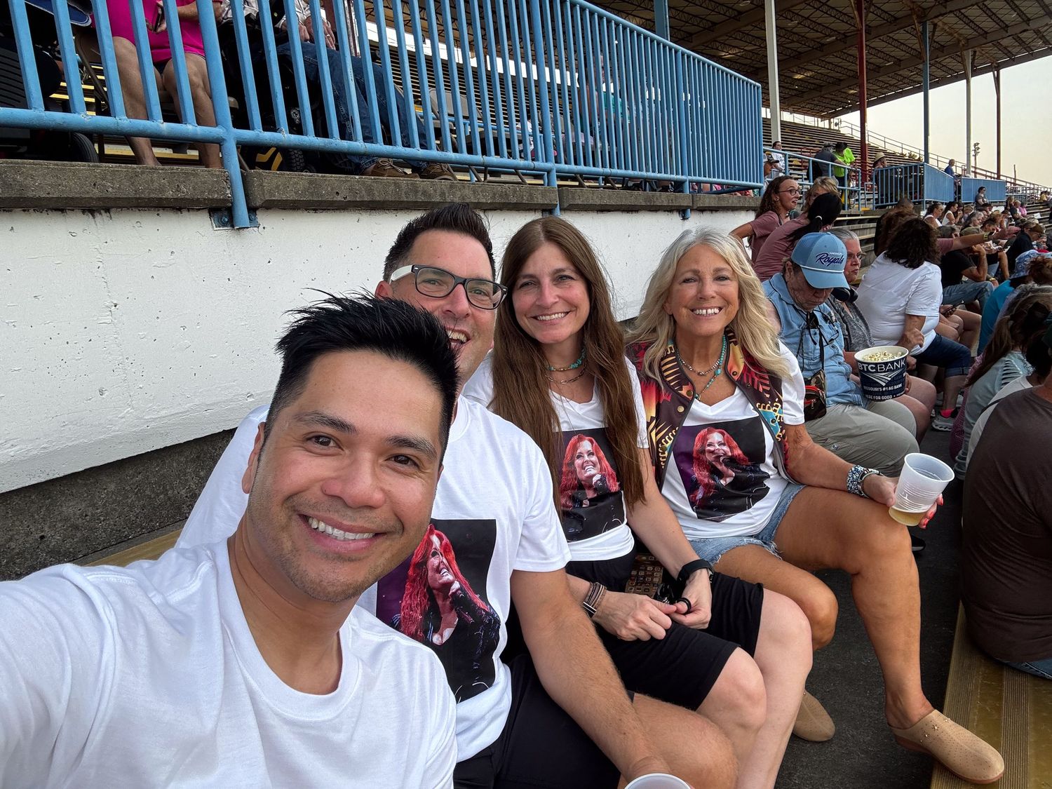 Four friends taking a selfie in stadium bleachers, wearing matching graphic t-shirts at an outdoor event.