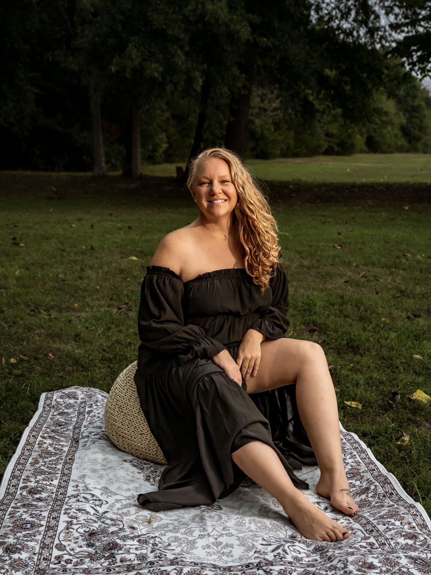 Woman in black off-shoulder dress sitting on blanket in park, smiling with curly blonde hair.