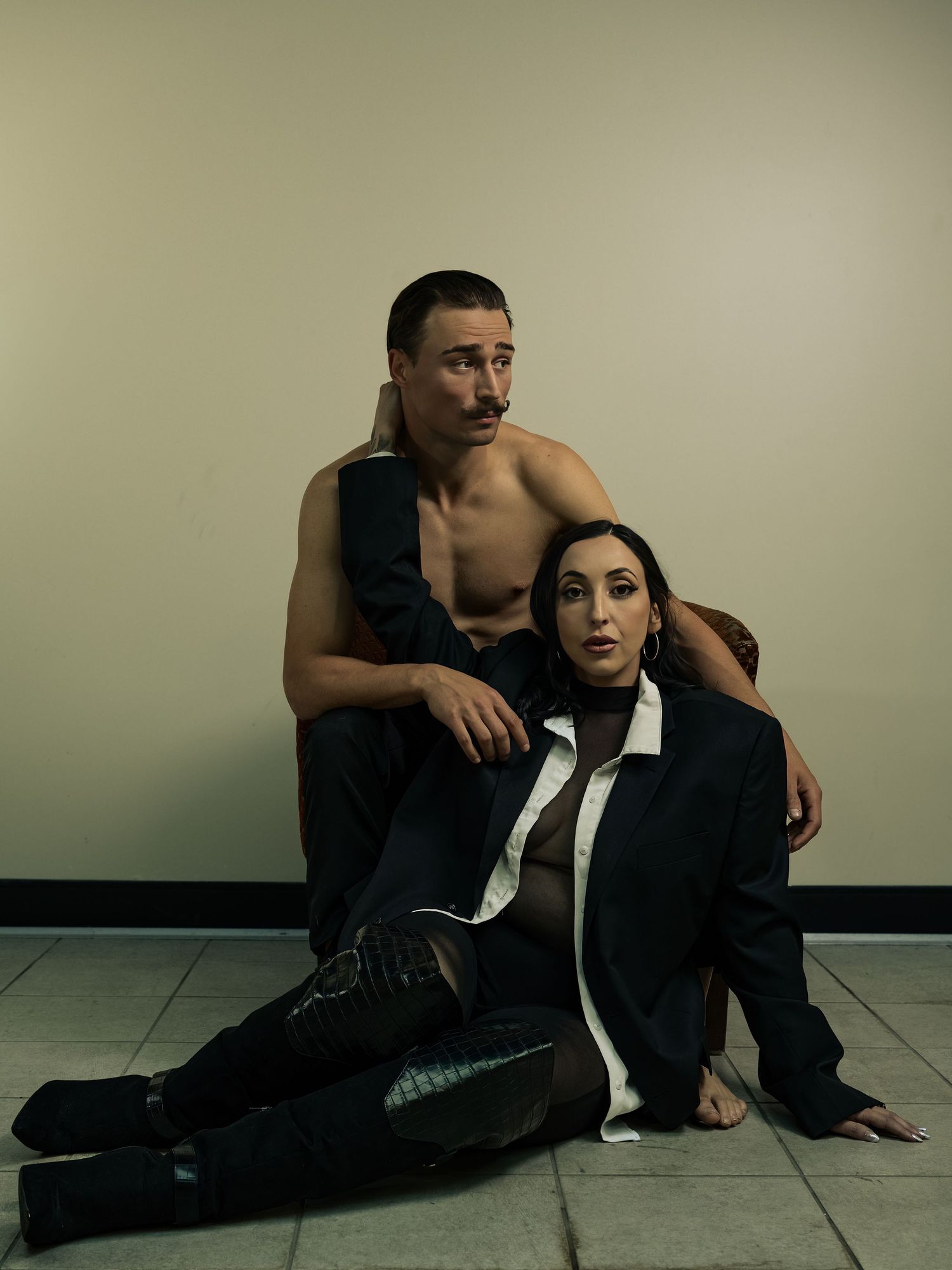 A shirtless man and a woman in a black blazer pose dramatically on a tiled floor in a moody editorial shoot.