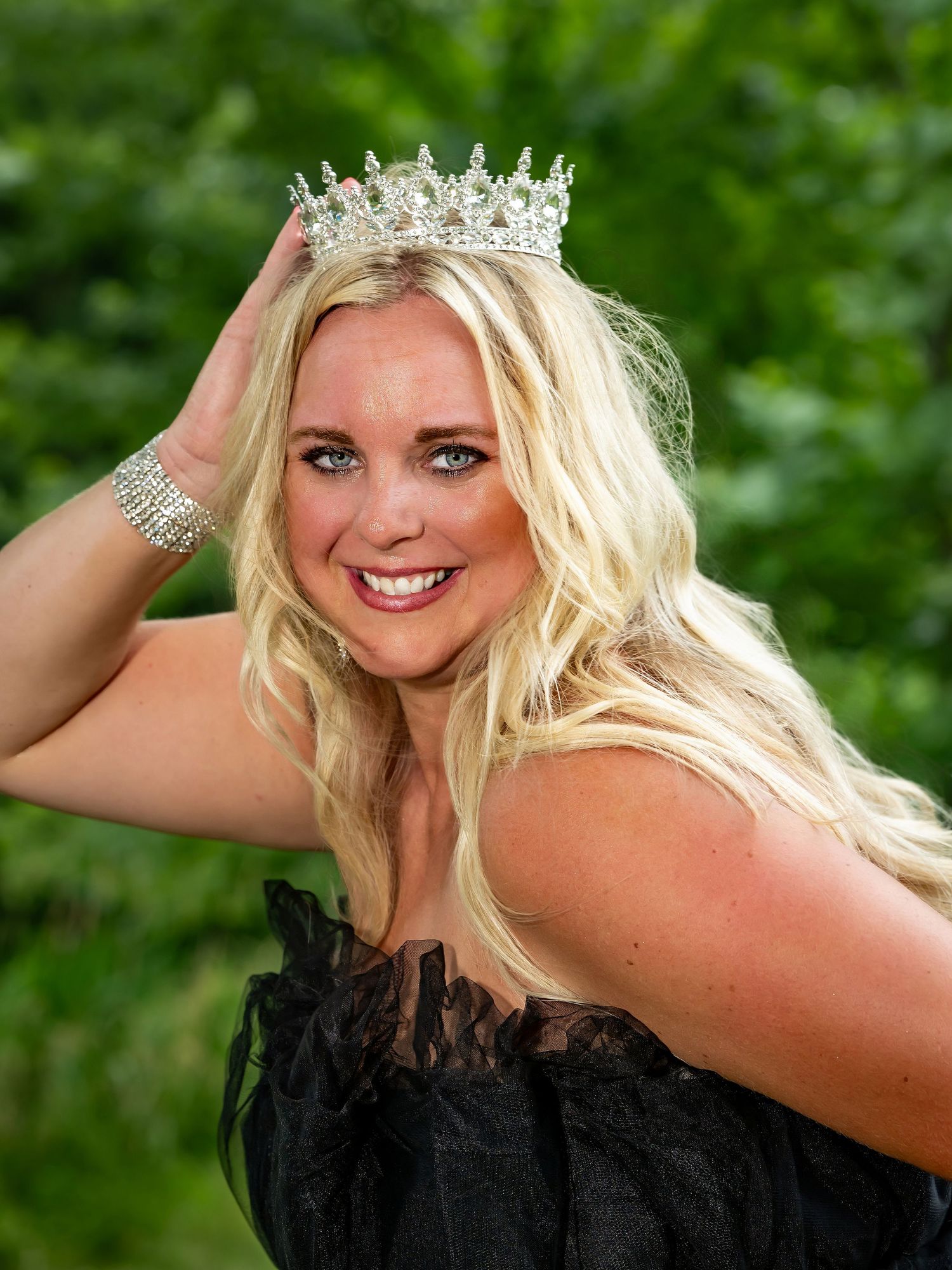 Person wearing a crown and black lace top poses against green foliage background.