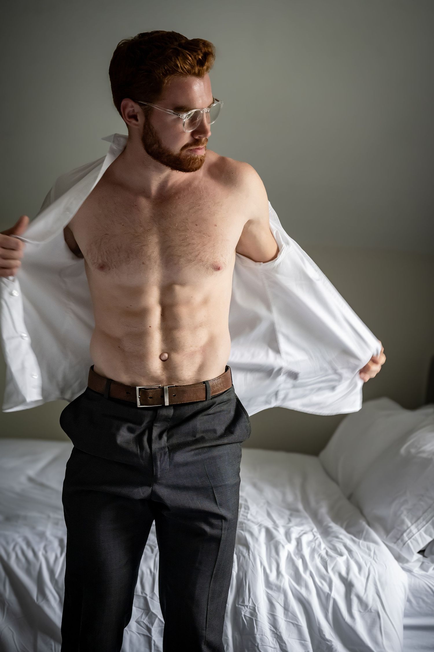 Redheaded bearded man removing white shirt revealing toned physique, wearing gray trousers and glasses in bedroom.