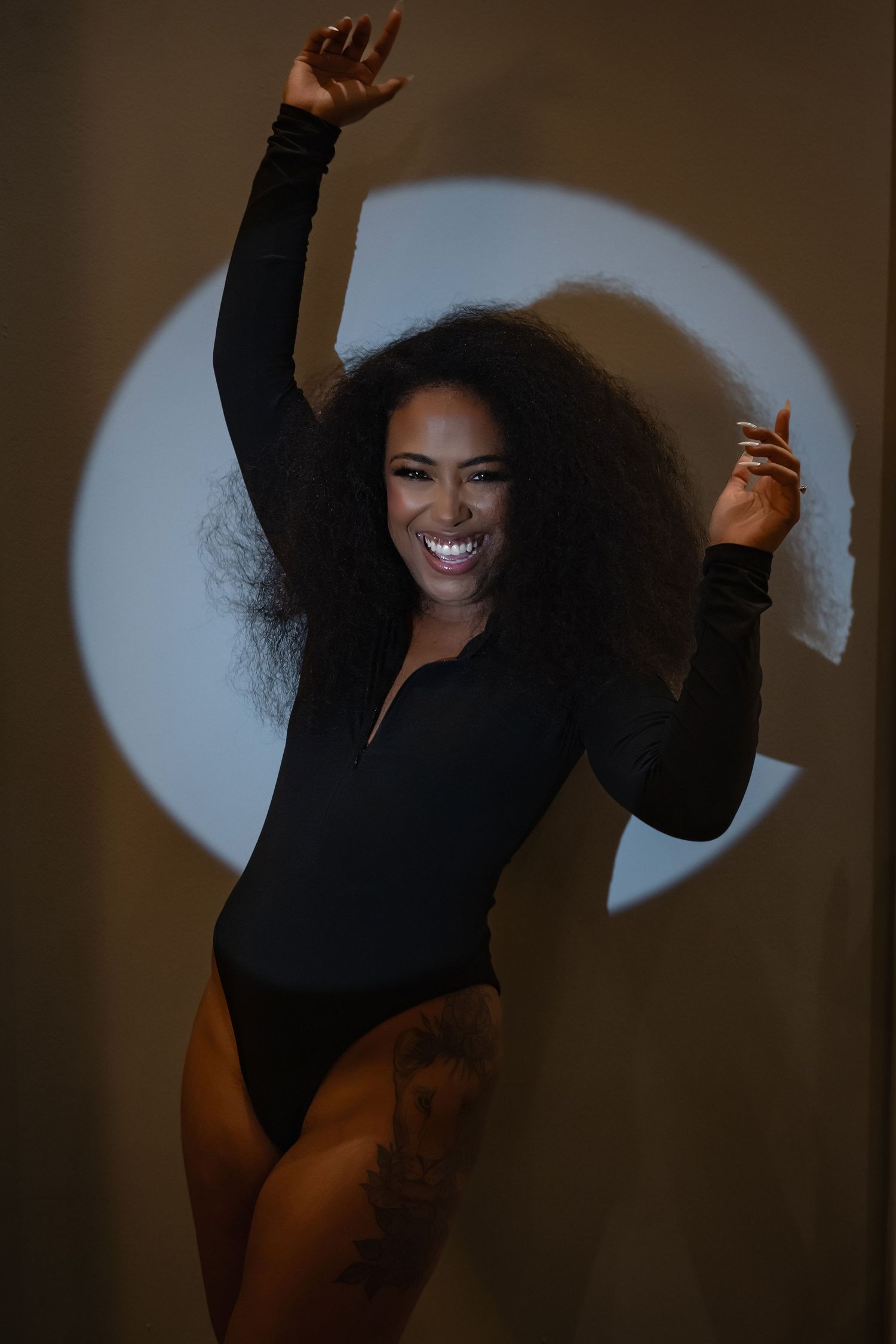 A joyful Black woman with curly hair dances in a black bodysuit, arms raised, smiling before a circular spotlight.