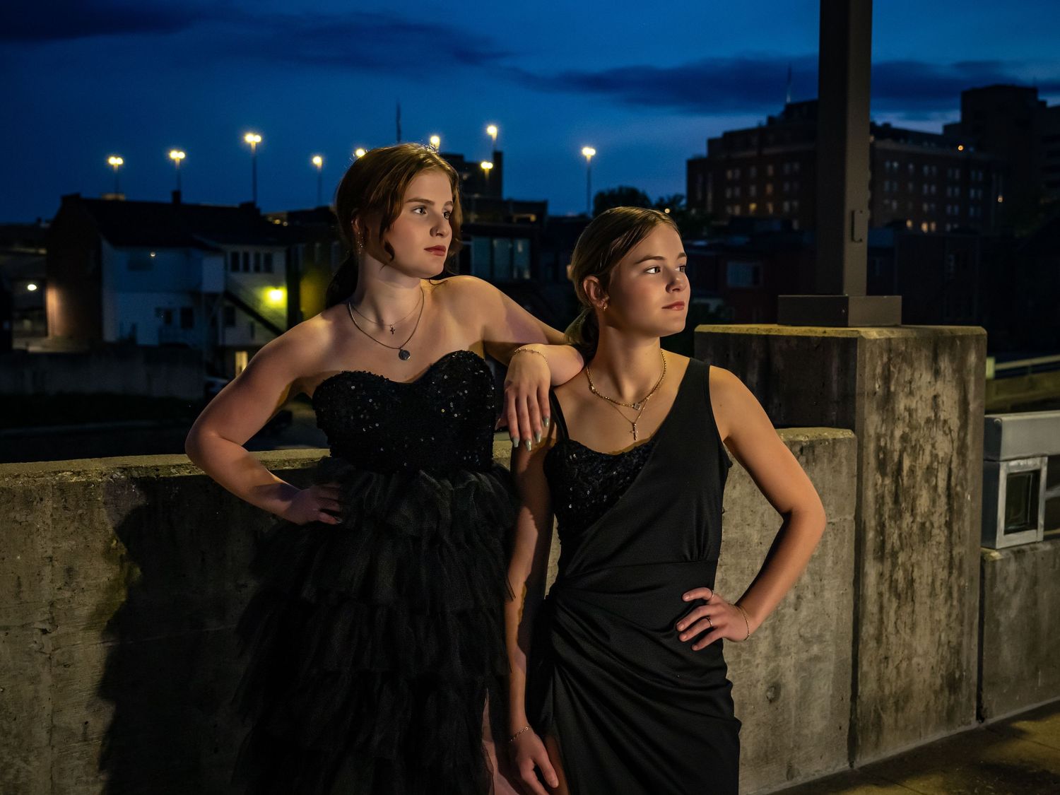 Two figures in formal attire pose on a rooftop at dusk with city lights glowing in the background.
