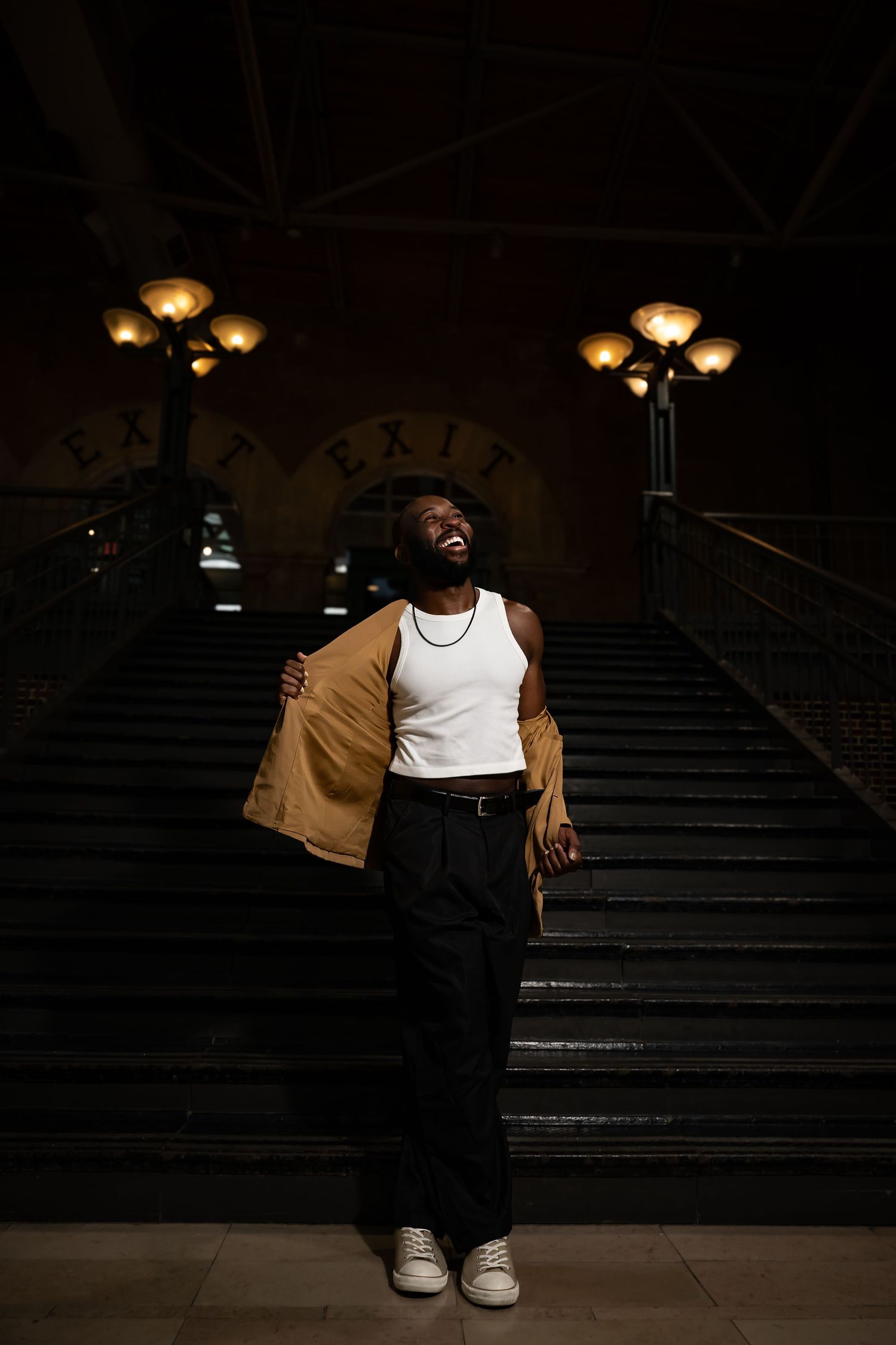 Expressive pose on dark stairs with ambient lighting creating dramatic shadows.