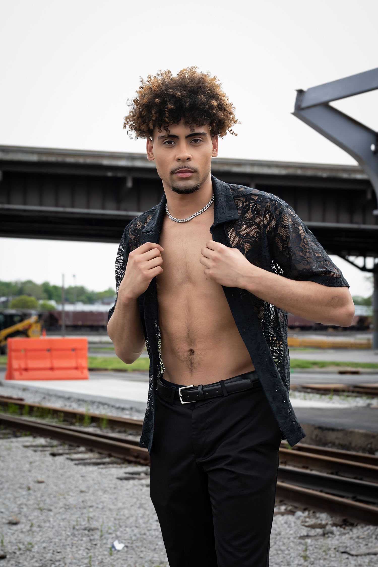 Fashion model poses shirtless in black lace shirt and pants near railroad tracks in urban setting.