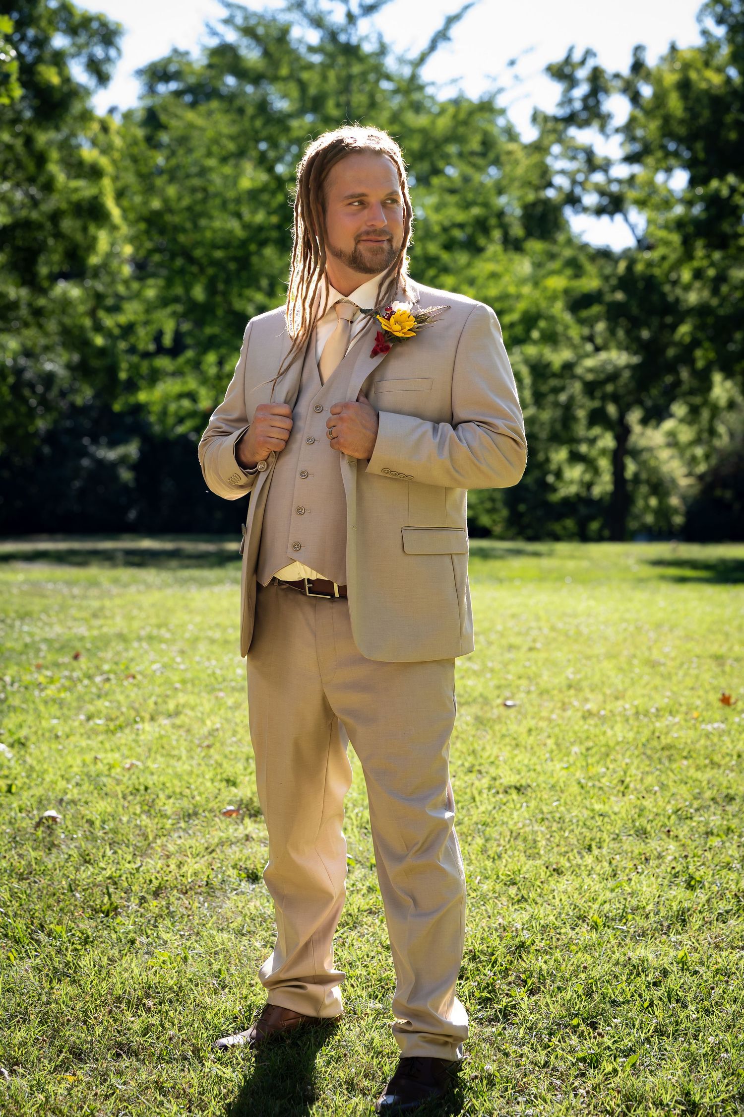 Groom in tan suit poses outdoors on sunny lawn with natural backdrop.