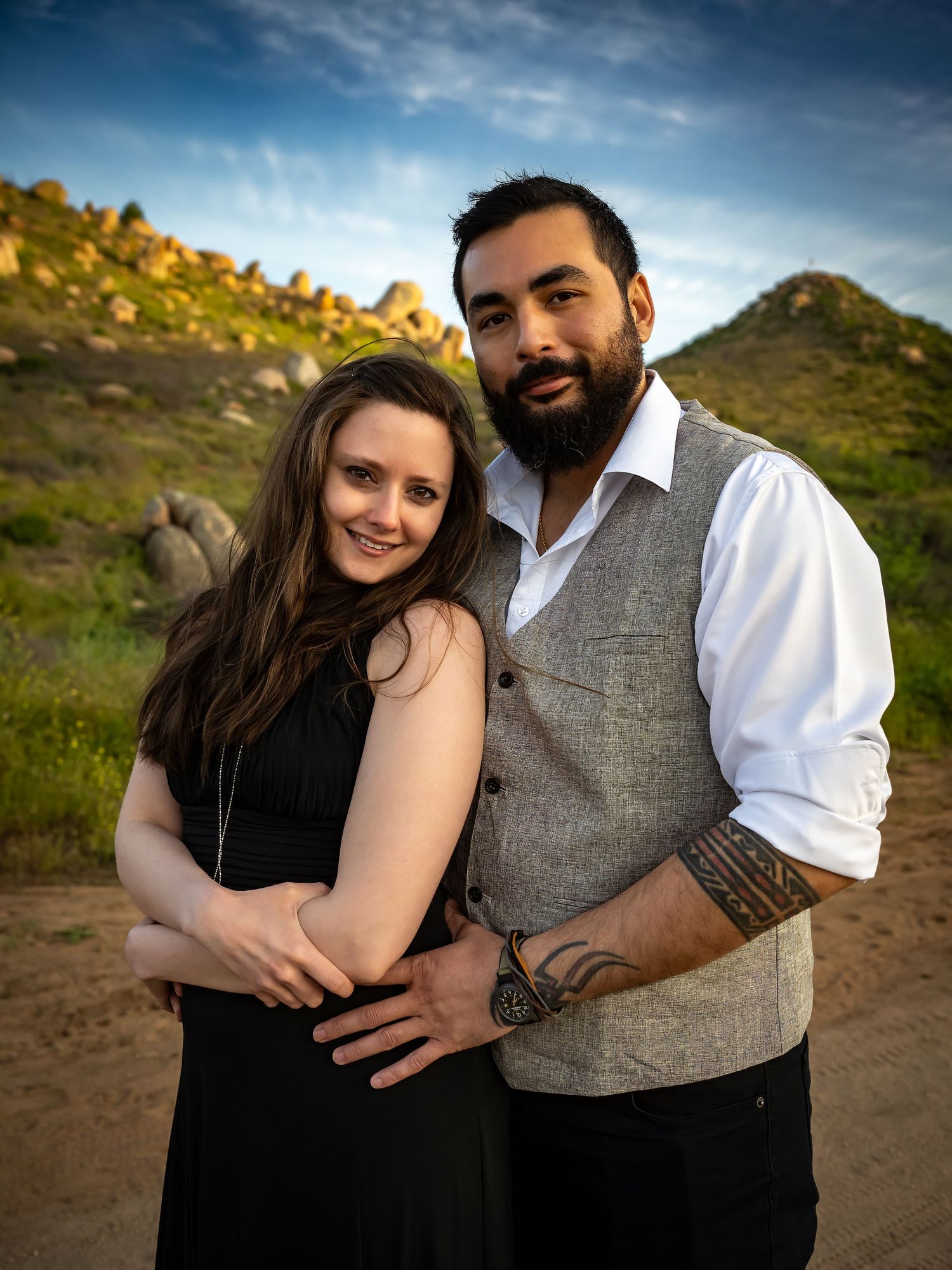 A couple embracing outdoors against a backdrop of mountains and blue sky.