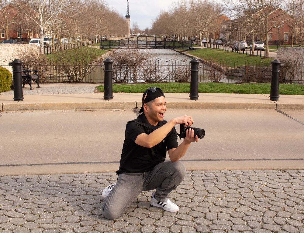 Photographer crouching outdoors near a canal, holding a camera and smiling, surrounded by trees and black bollards.