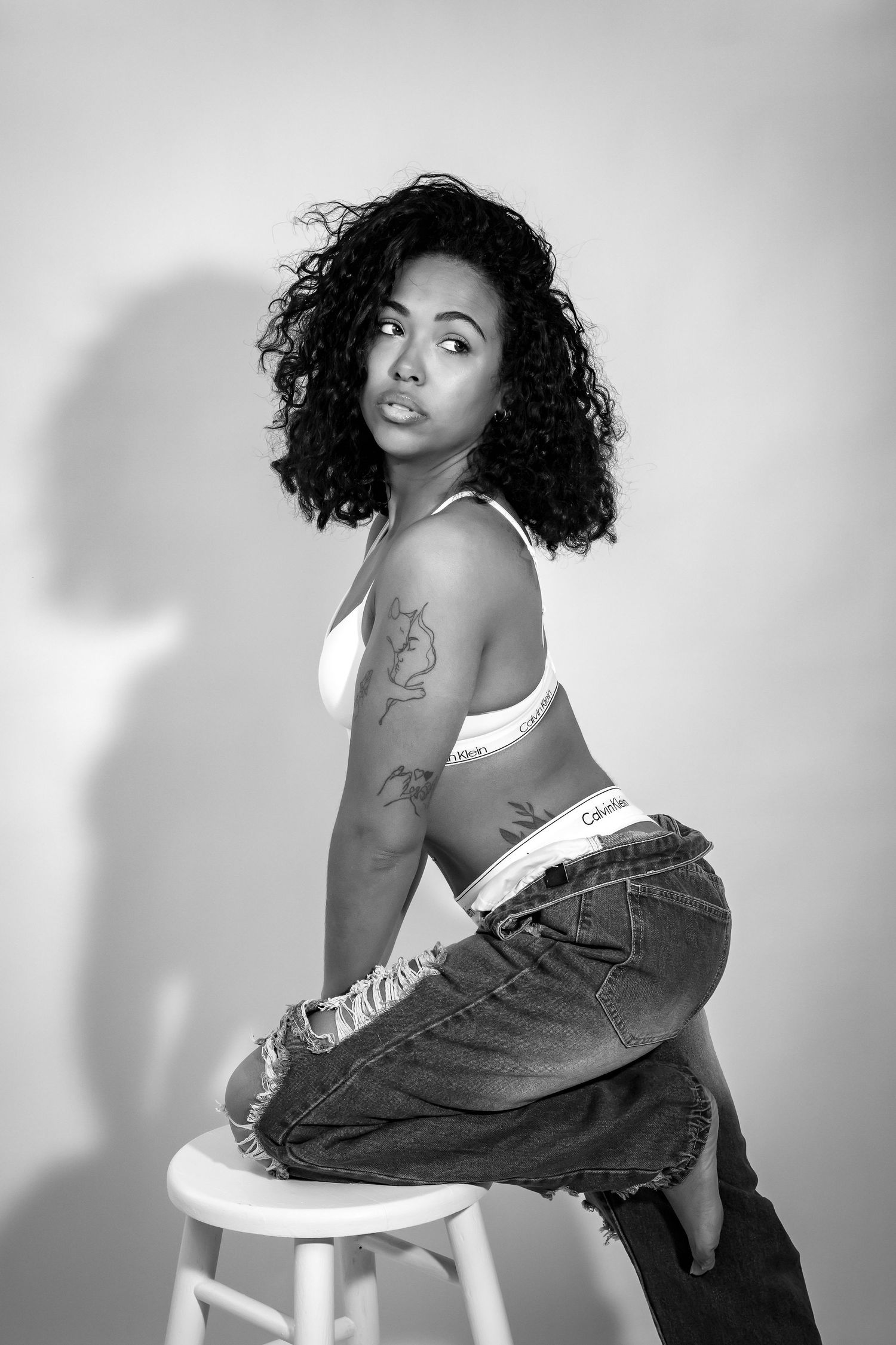 Black and white studio portrait of model in casual denim and white top sitting on stool.