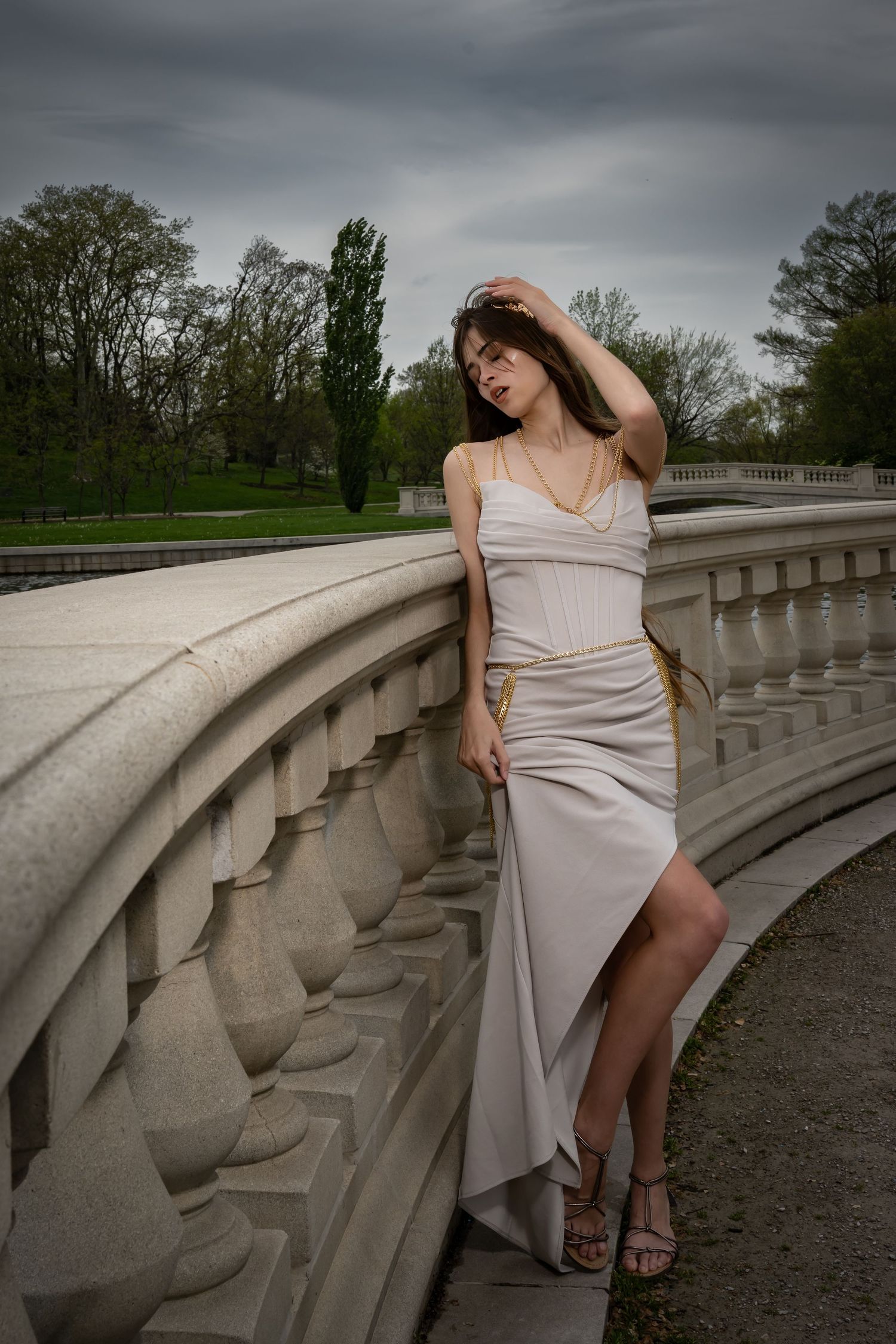 Figure in a cream dress poses dramatically on a curved stone balustrade under an overcast sky.