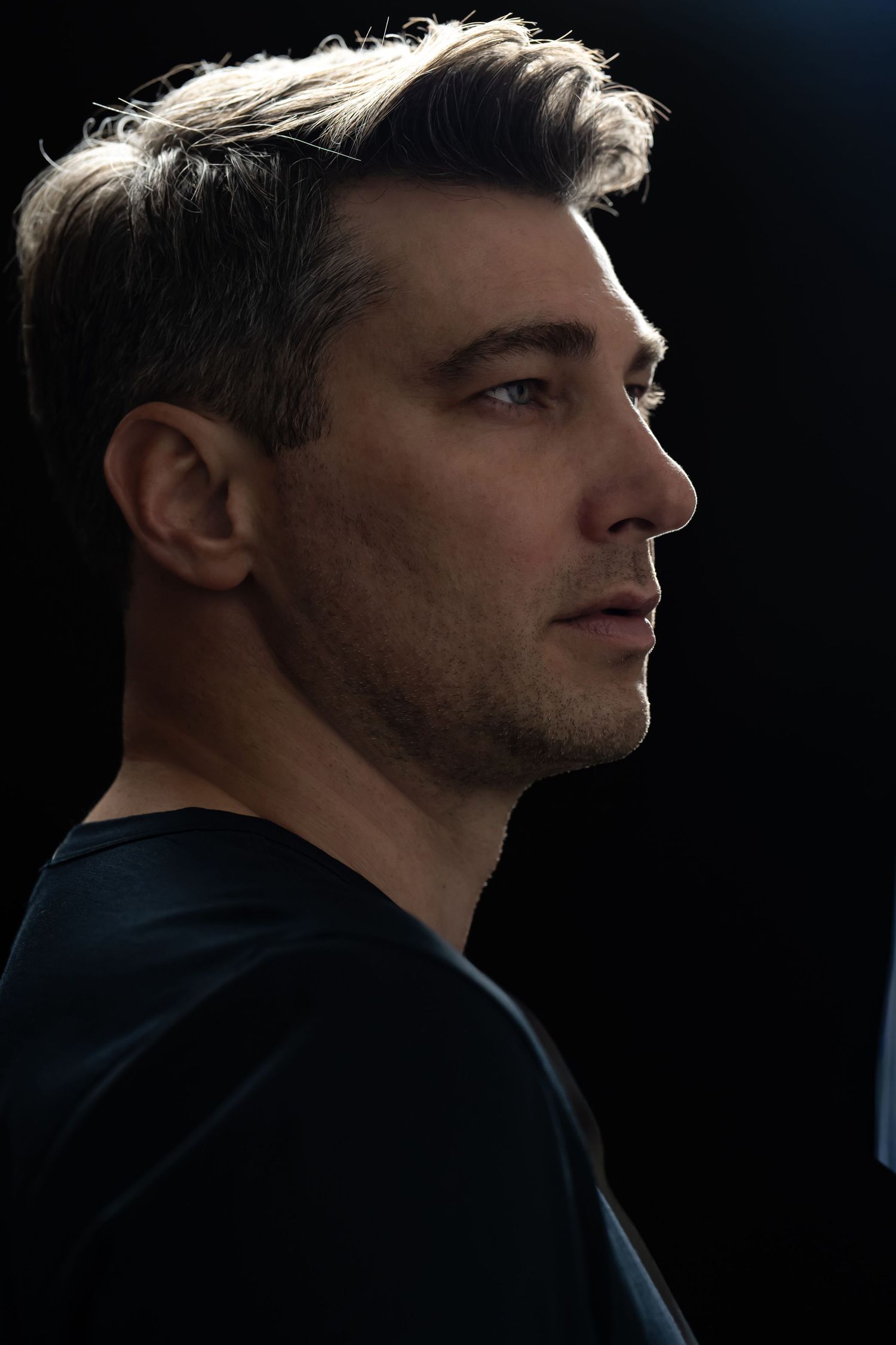 Side profile portrait of a man with salt-and-pepper hair in a black shirt against a dark background.