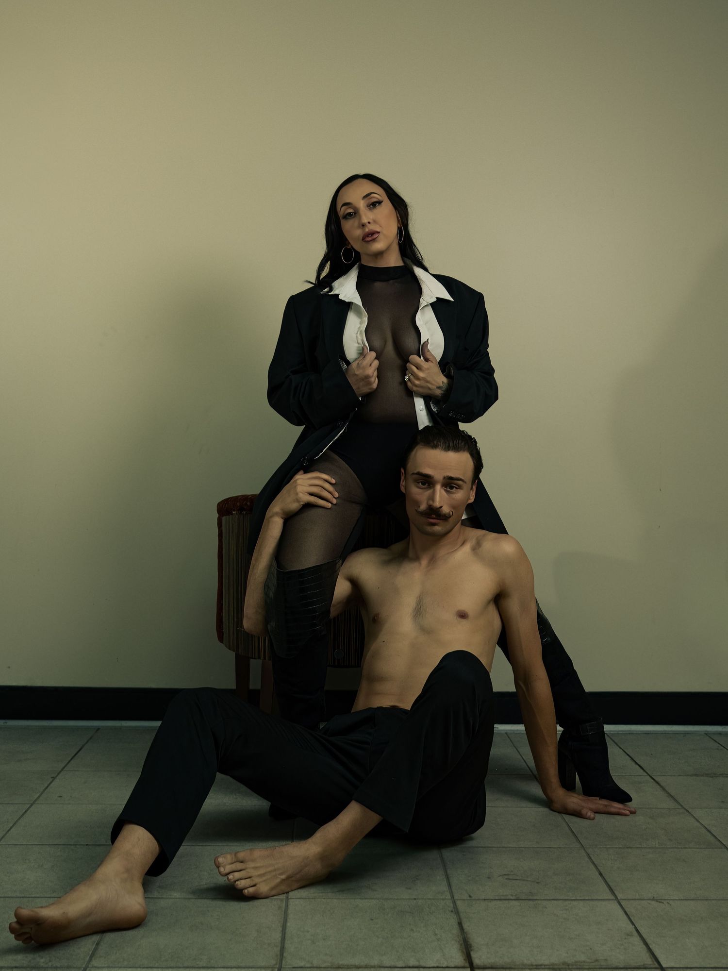 A woman in a black blazer sits on a chair above a shirtless man in black pants on a tiled floor.