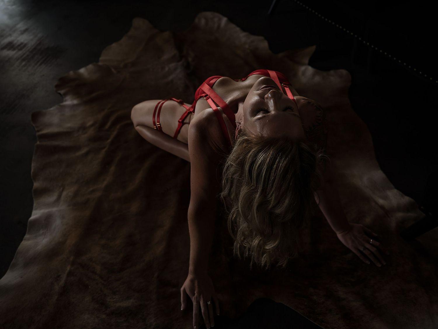 Woman in red lingerie posing dramatically on a fur rug in a dark, moody boudoir photography setting.