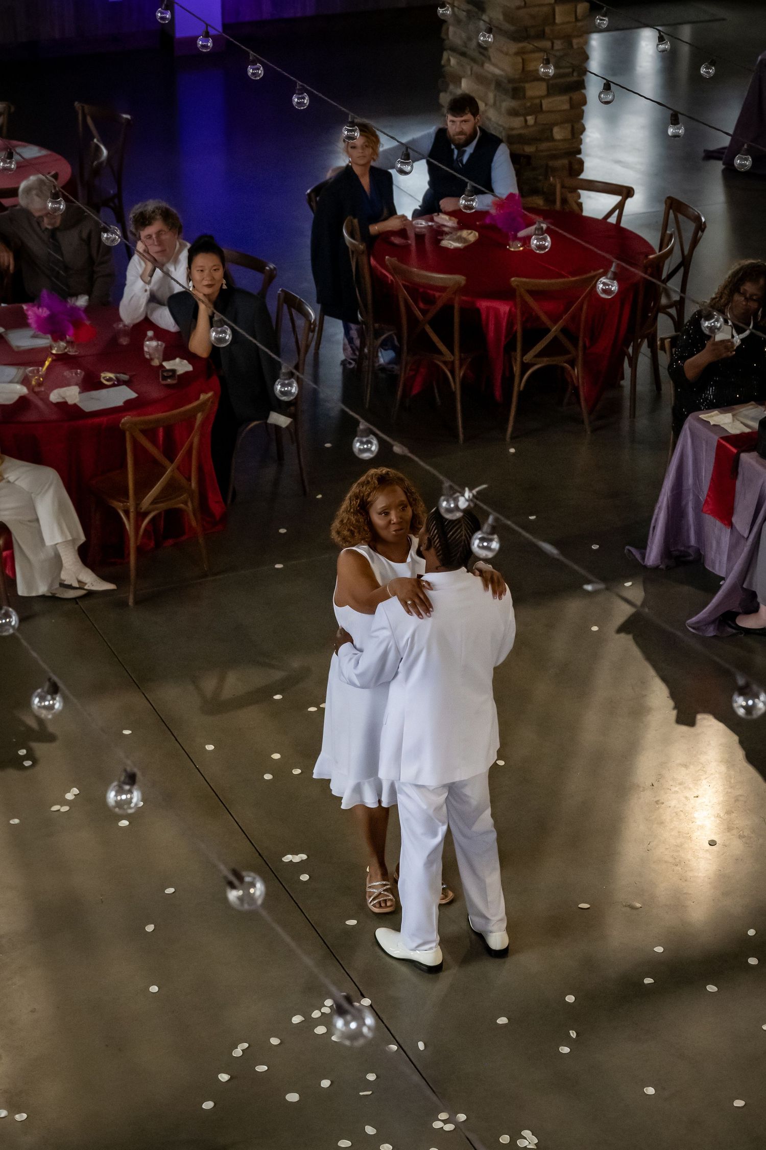 Wedding reception dance floor with guests in white attire and red tables in background.