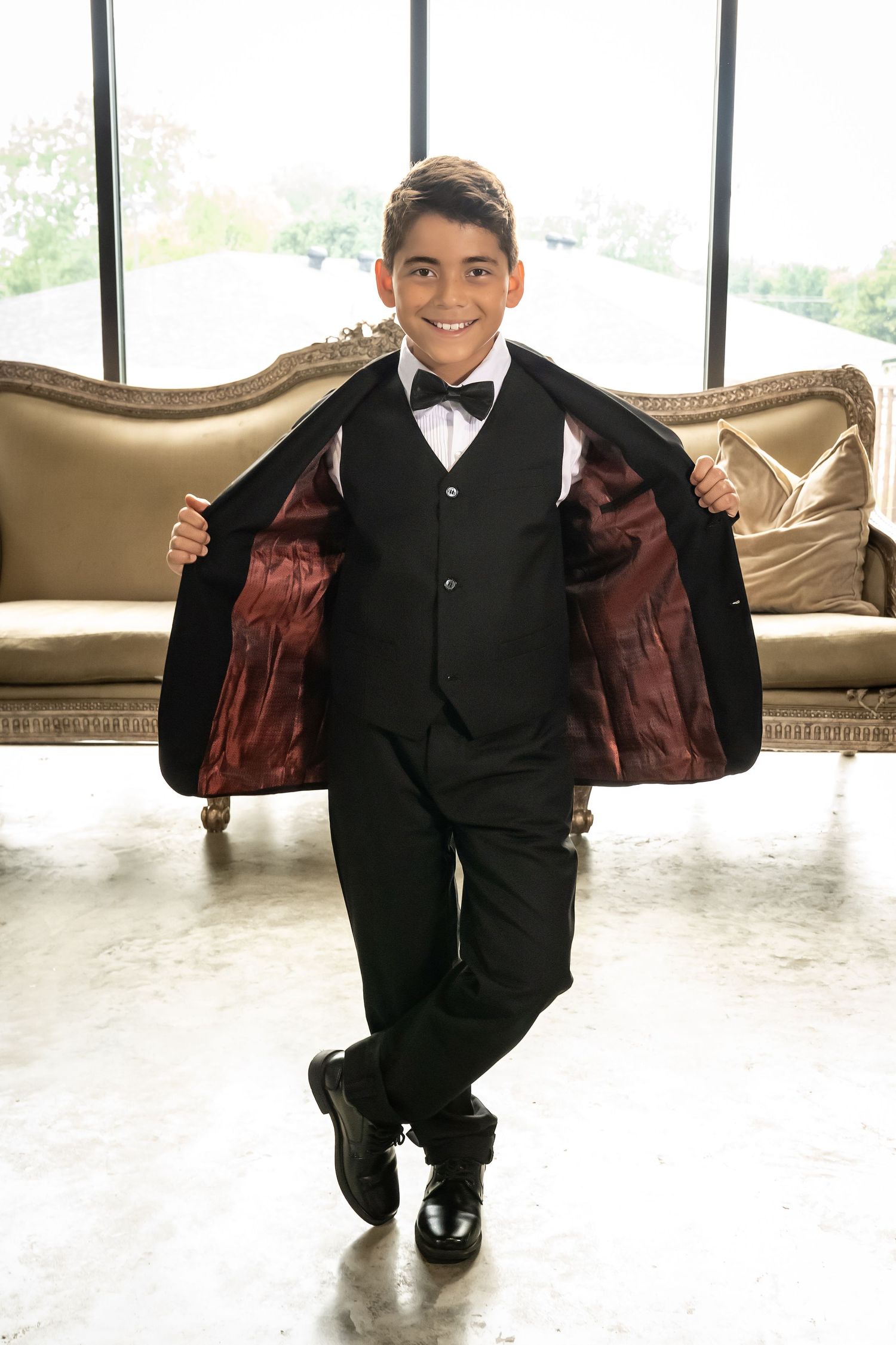 Young ring bearer in black suit playfully posing on beige couch.