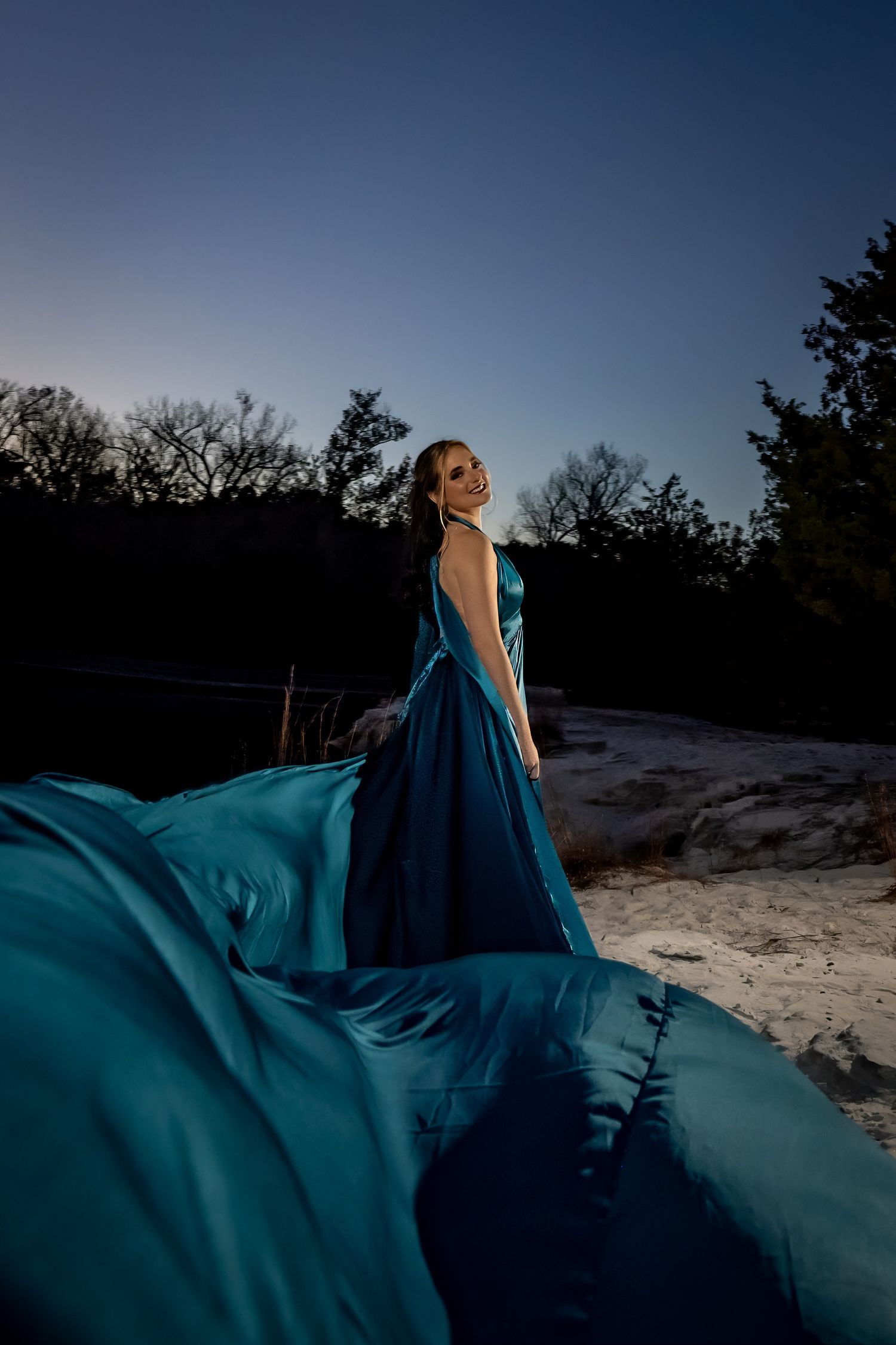 Figure in flowing blue dress poses dramatically on sandy beach at dusk.