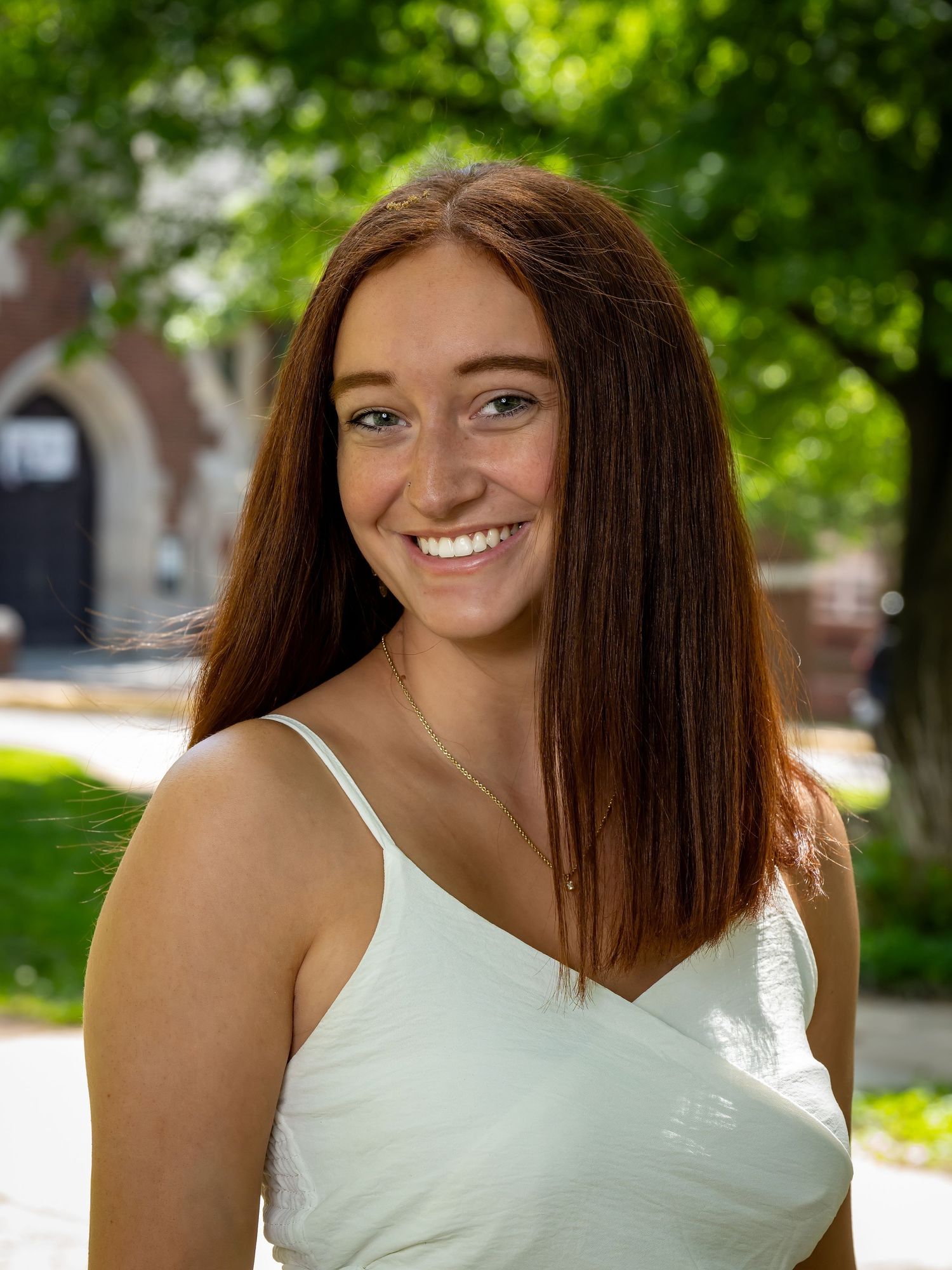 Graduate in white dress poses on sunny campus lawn.
