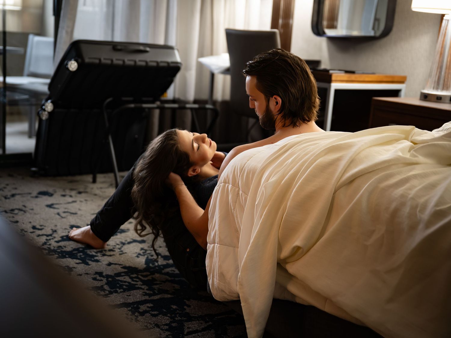 A couple in wedding attire lies on a white bed in a romantic pose during a photoshoot.