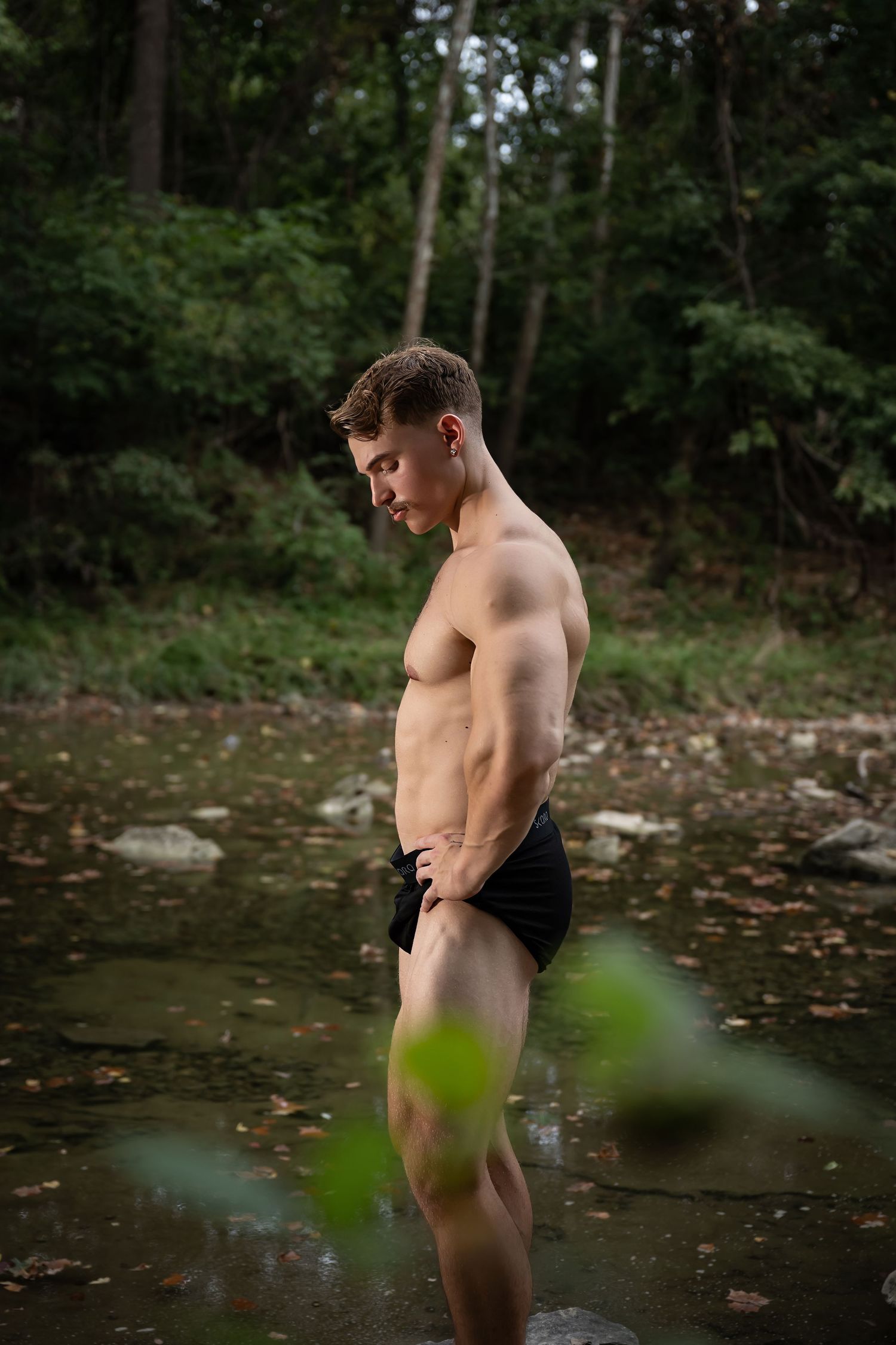 Muscular shirtless man standing in a shallow forest stream wearing black shorts, surrounded by lush green trees.