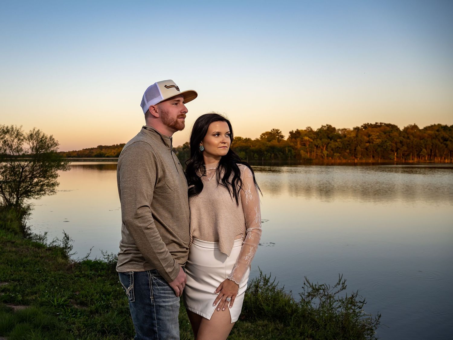 Couple standing by serene lake during sunset with calm water reflections.