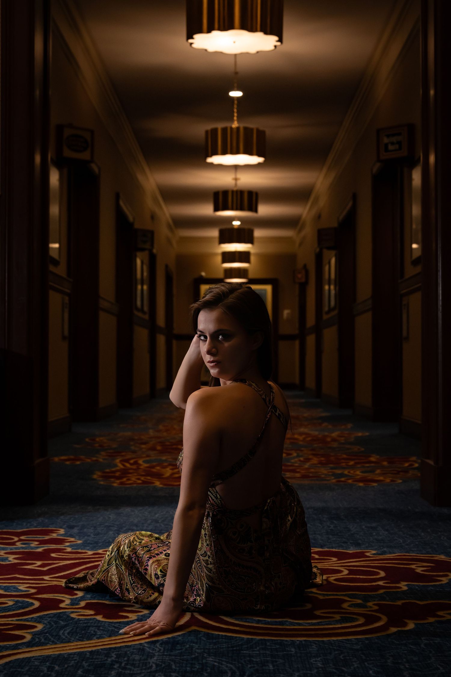 Moody portrait of model sitting on patterned carpet in dark hotel corridor.