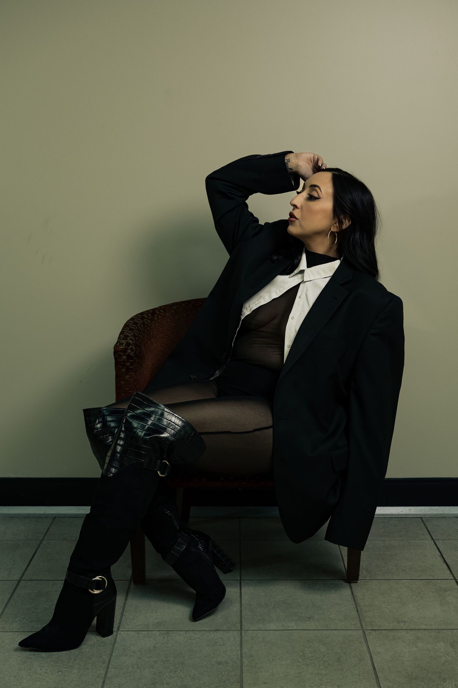 Woman in black blazer and knee-high boots poses elegantly in leather chair against neutral wall.