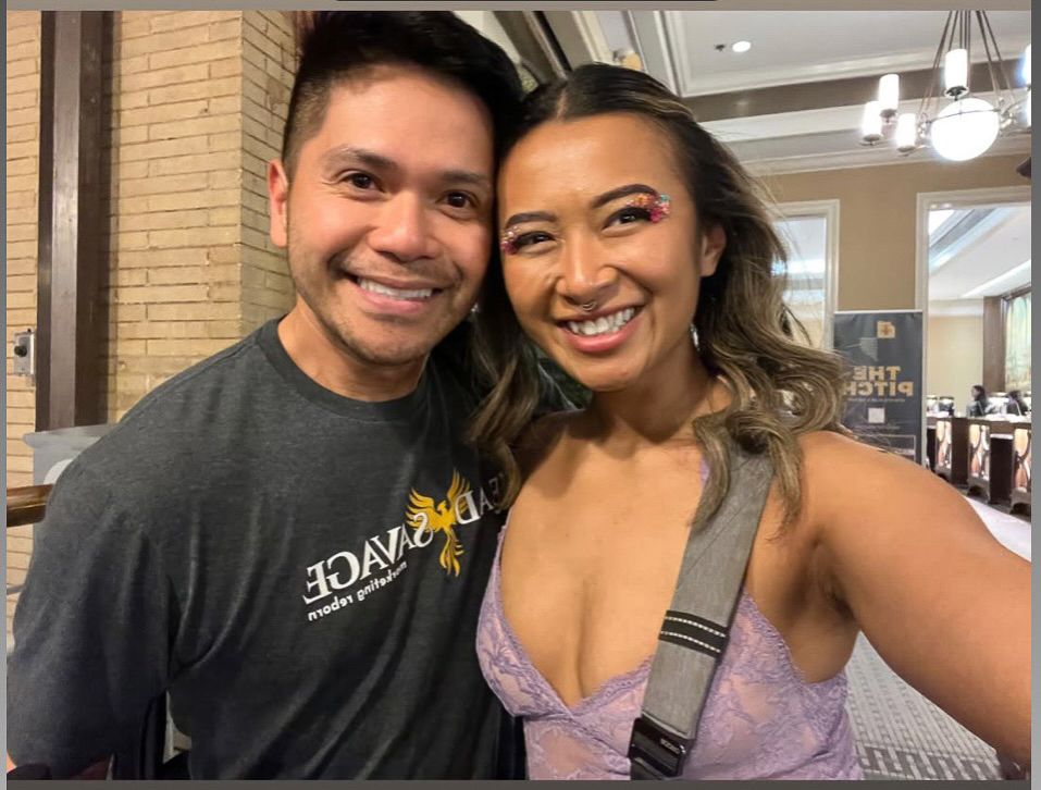 A couple smiles together in a casual indoor setting, with one wearing a black t-shirt and the other in purple.