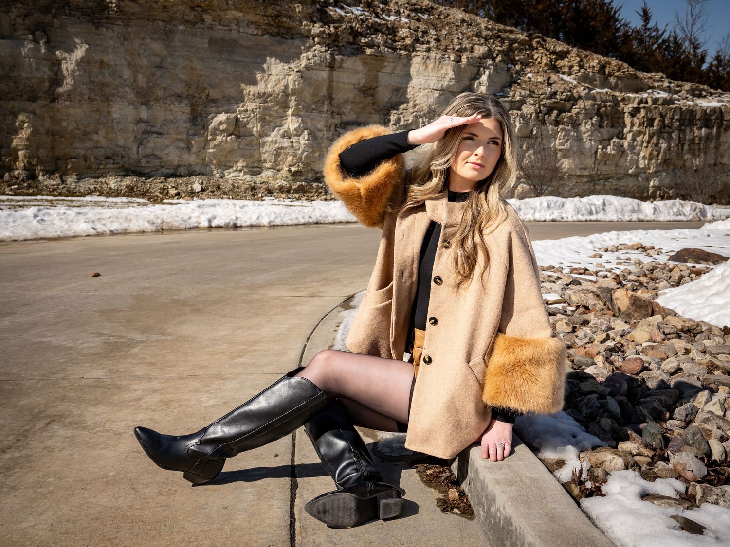 Model in tan coat poses outdoors against stone wall in snowy winter setting.
