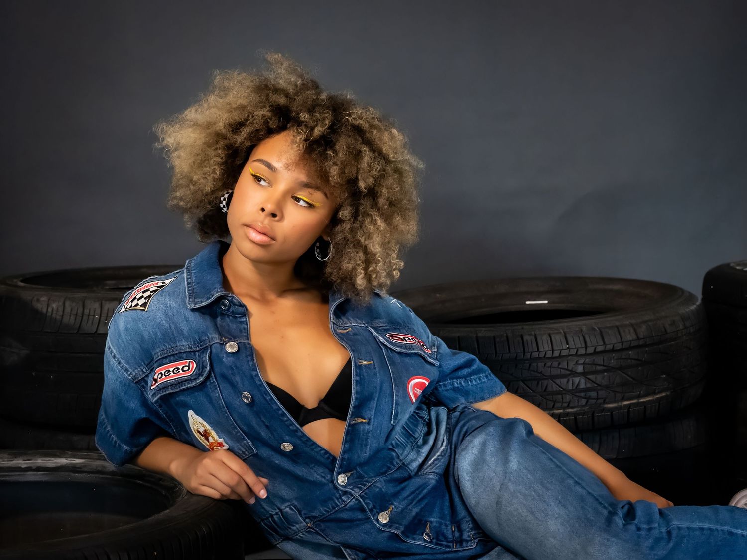 Fashion portrait with denim jacket and tires in moody lighting.