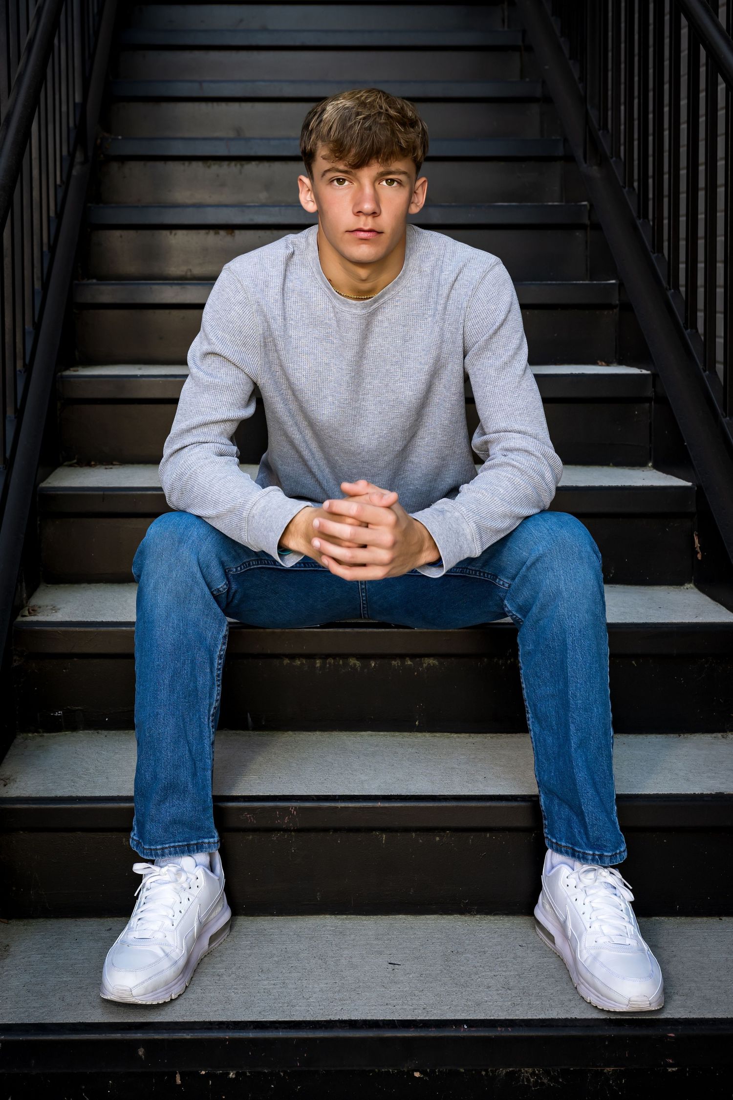 Person in gray sweatshirt and jeans sits on dark stairs wearing white sneakers in moody lighting.