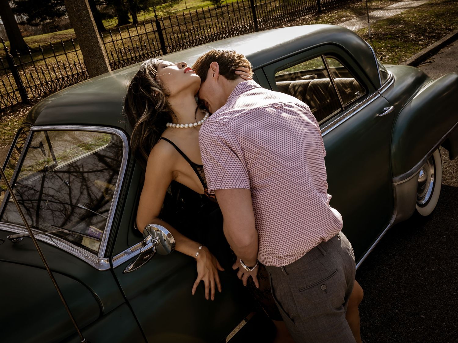 Couple sharing a romantic kiss leaning against a vintage classic car outdoors in soft natural light.
