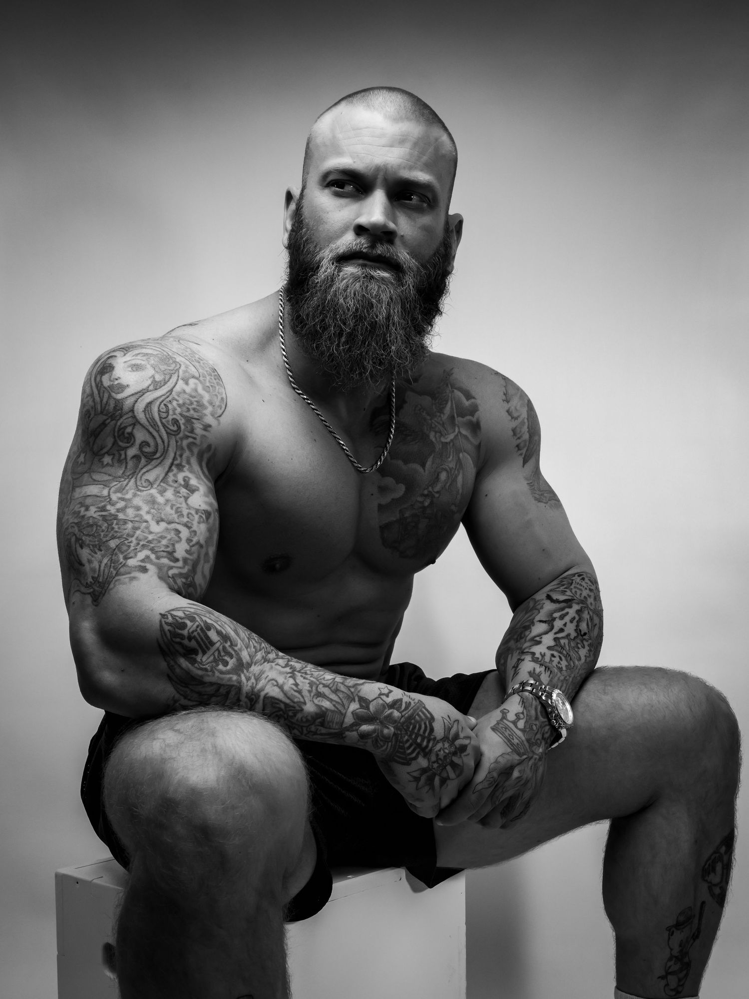 Black and white photo of a muscular bald man with a full beard and extensive tattoos sitting shirtless.