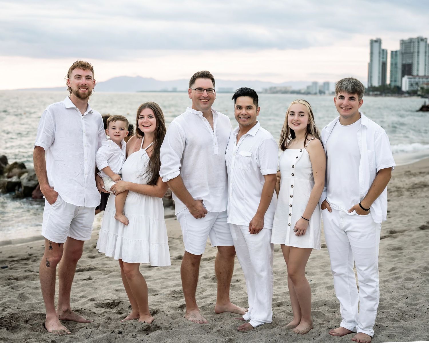 Beach family photo with everyone dressed in white outfits near ocean.