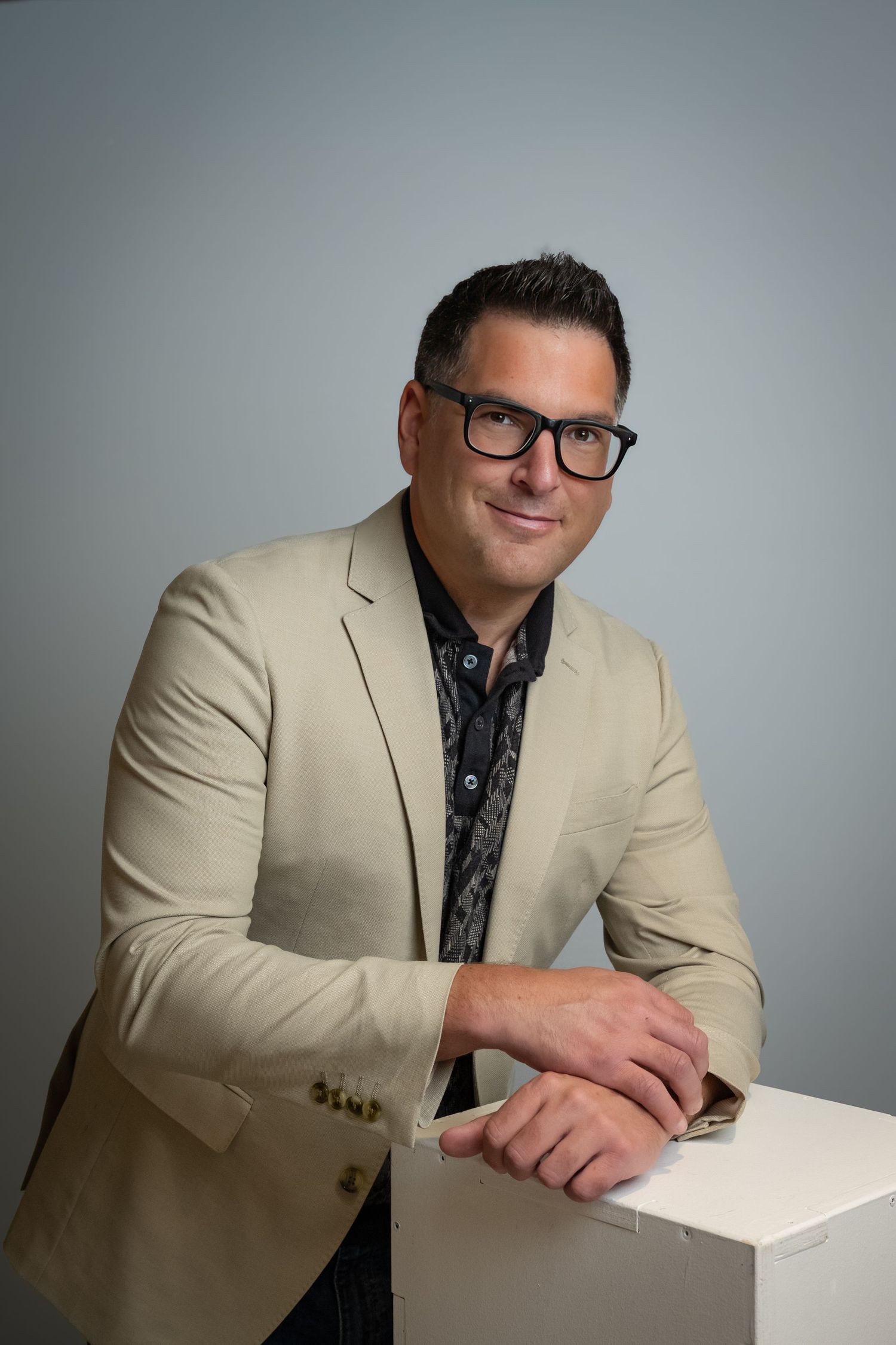 Man in beige blazer and black glasses leaning on white cube, smiling in professional studio portrait.
