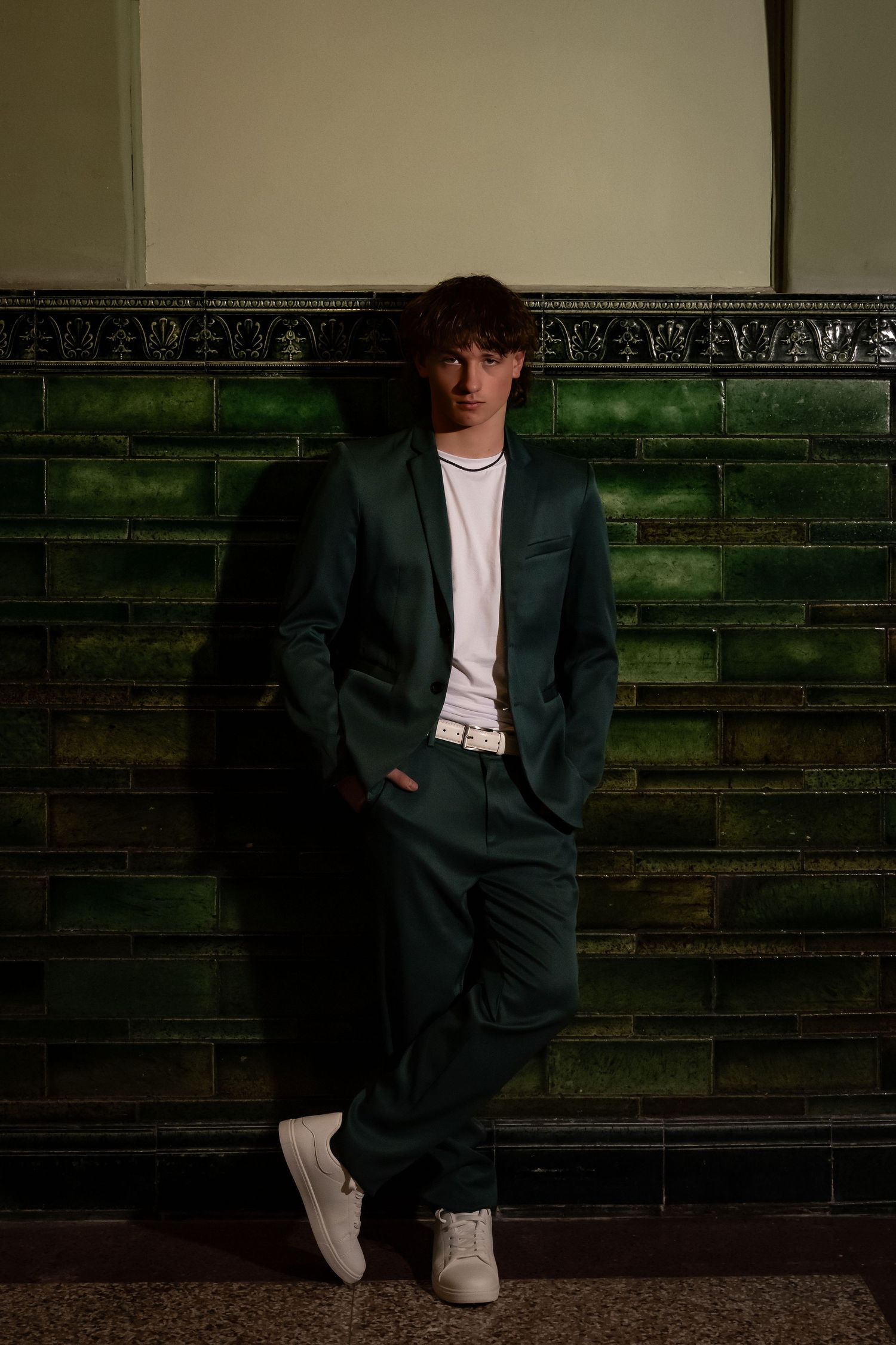 Moody portrait of person in dark green suit against tiled wall.