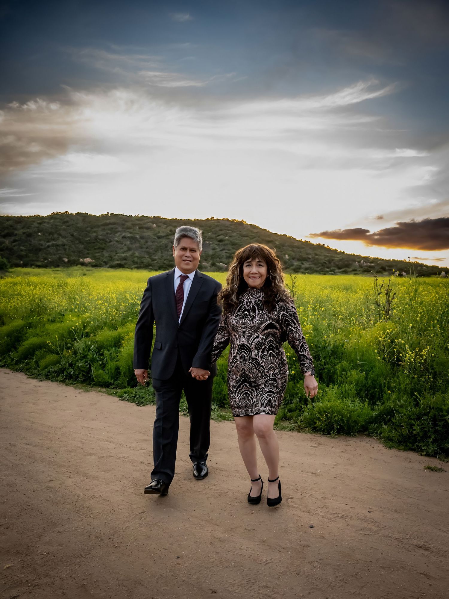 A couple poses on a dirt path with yellow wildflowers and mountains in the background.