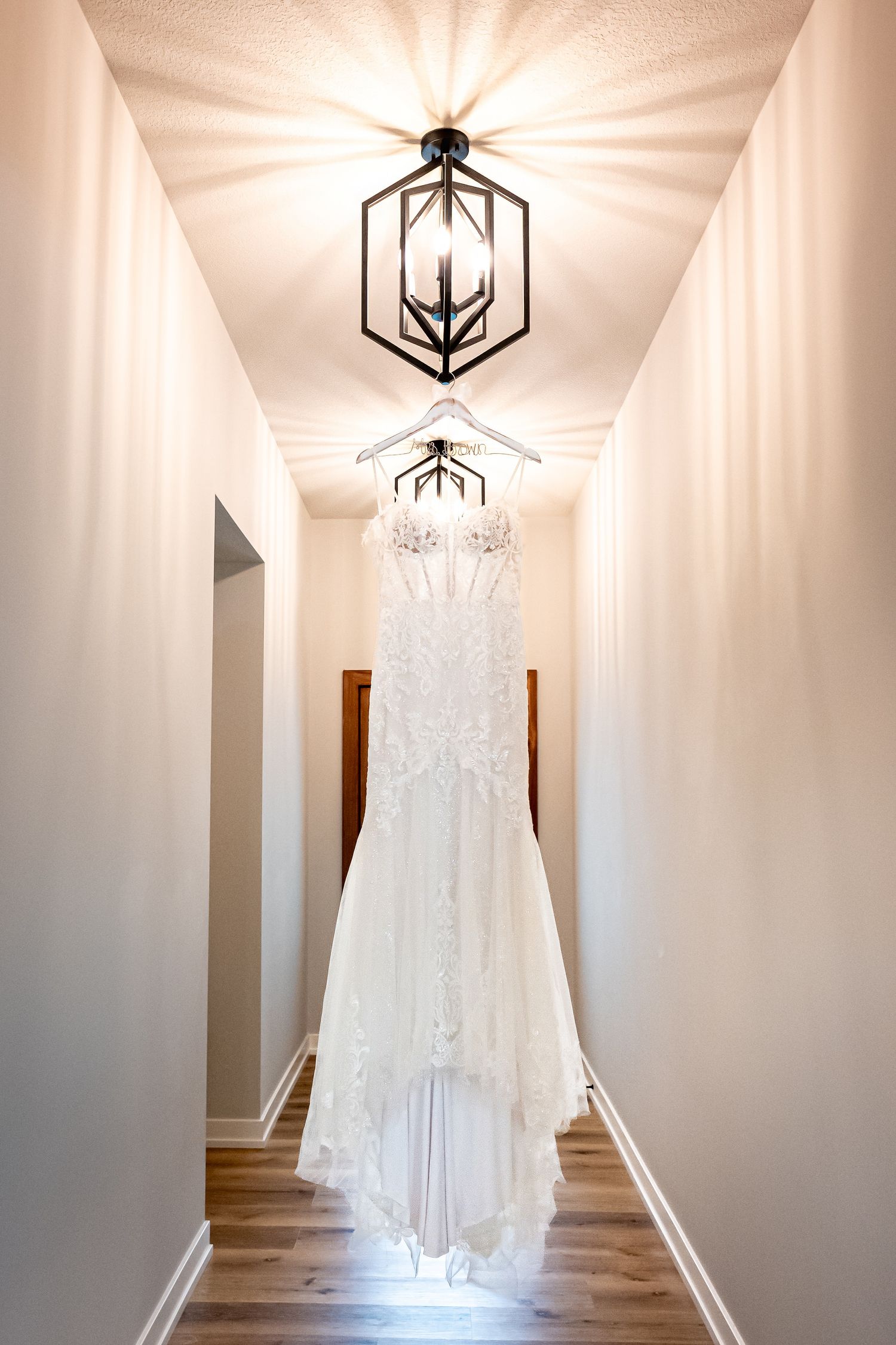 Elegant white lace wedding dress hangs in bright hallway with modern geometric light fixtures.