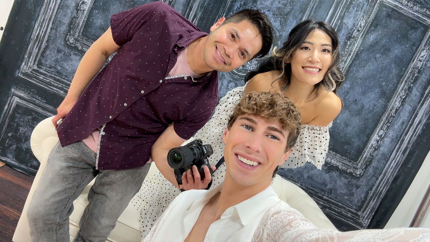 Three friends pose for a photo in a studio, with one holding a camera, against a decorative dark door backdrop.