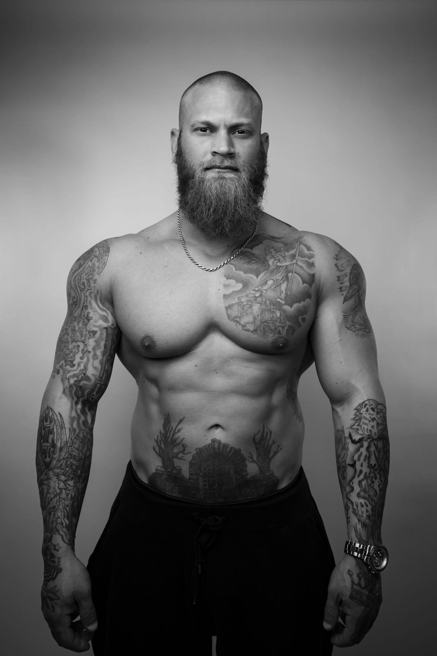 Black and white portrait of a muscular bald man with a beard and tattoos posing shirtless in a studio.